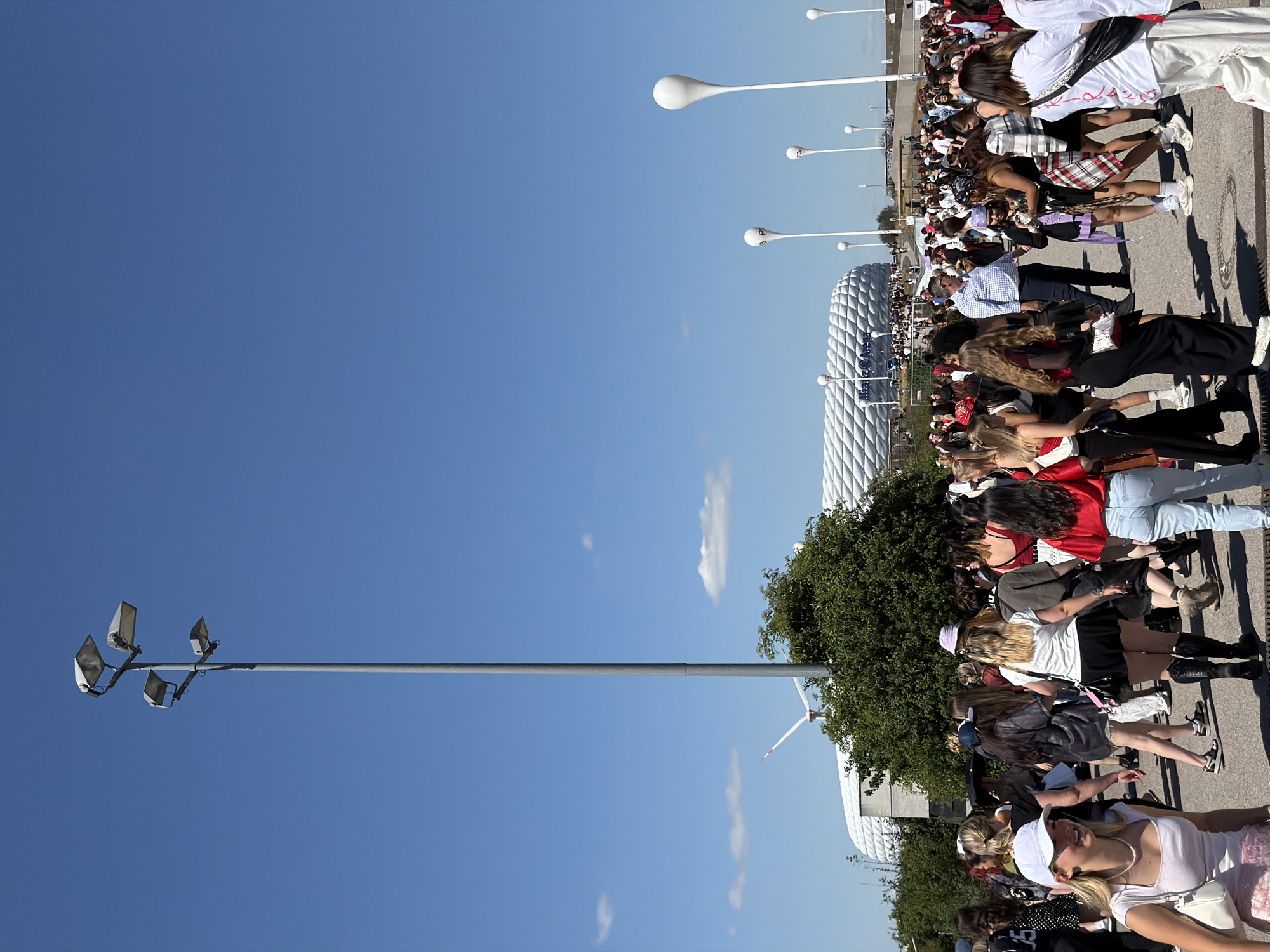 In Munich, Germany, a large crowd of people, predominantly young women, are gathered outdoors under a clear blue sky. Many are dressed casually, with some wearing festival-like attire, including hats and distinctive hairstyles. They are walking on a paved area, with a stadium in the background that appears to be the Allianz Arena, identifiable by its unique illuminated facade. Several tall, slender light poles with spherical or teardrop-shaped lamps are positioned throughout the scene, along with larger stadium lighting poles. A wind turbine is visible in the distance, suggesting the location is an open area. The atmosphere appears to be one of a public gathering or event, with people moving and socializing.