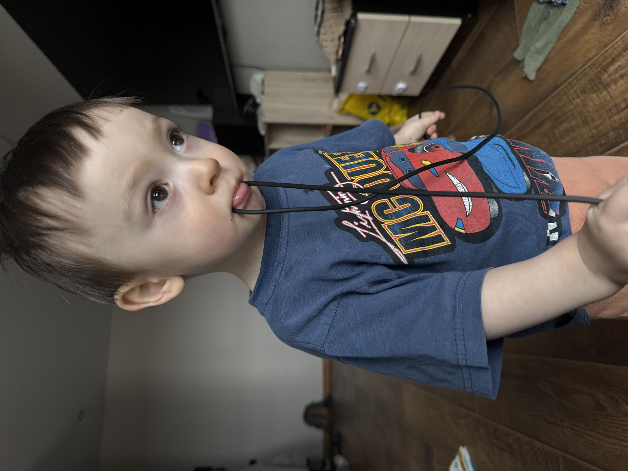 A child is holding a black electrical cord in their mouth. They are wearing a blue t-shirt with a red cartoon car design. The child is looking upwards and to the left. The background shows a wooden floor and some furniture. There is no indication of Izhevsk, Russia.
