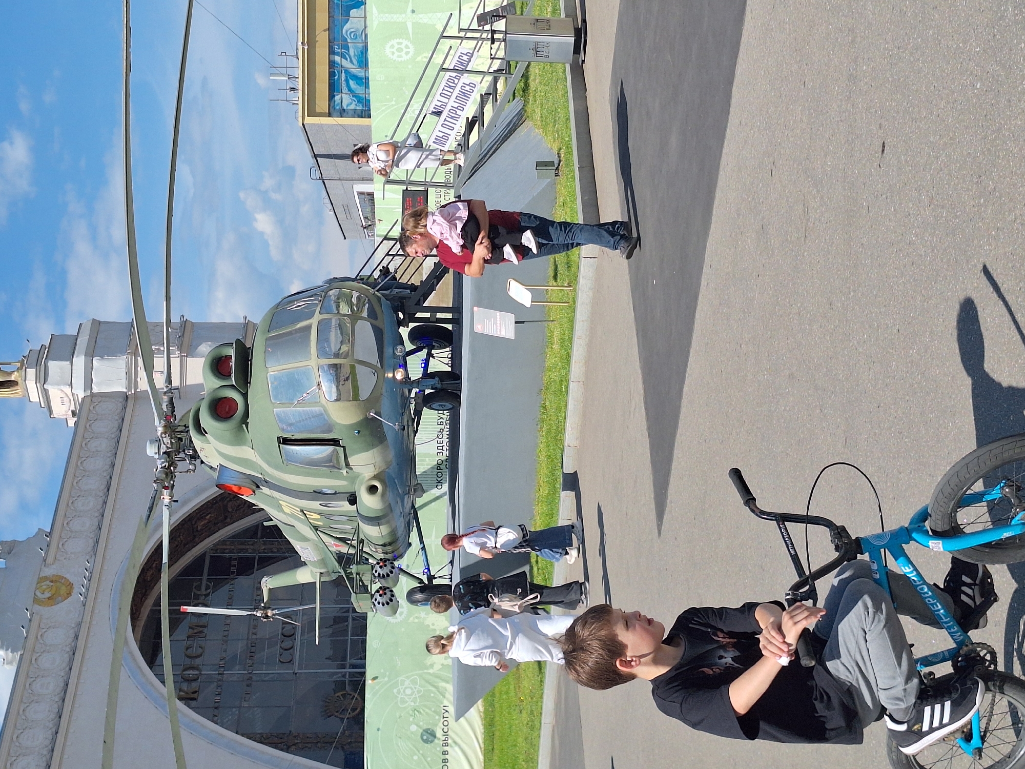 This daytime image, set in Moscow, Russia, captures an outdoor scene, likely at the VDNKh exhibition grounds, under a clear blue sky with scattered clouds.
In the foreground, a young boy with brown hair, dressed in a black t-shirt and grey pants, sits relaxed on a blue BMX bicycle, which has "WETHEPEOPLE" visible on its frame. He looks towards the left, with his hands resting on the handlebars. His shadow is cast on the paved ground next to the bike.
The central focus includes a large, light green military-style helicopter, resembling a Mil Mi-2, displayed on a platform. It sits adjacent to a grand building with ornate white facades, where the Cyrillic words "КОСМОС" (Kosmos/Space) and "CCCP" (USSR) are visibly inscribed, indicating the Cosmonautics and Aviation Center at VDNKh.
Several individuals and families are scattered throughout the mid-ground. A man wearing a red shirt is seen holding a small child. Further back, another woman stands near a set of stairs. Green banners are displayed, one repeating "МЫ ОТКРЫЛИСЬ" (We are open), while another partially reads "СКОРО ЗДЕСЬ БУДЕТ..." (Soon there will be...) and "В ВЫСОТУ!" (Up to height!). The scene depicts a casual, pleasant day with visitors exploring the various exhibits and attractions of the complex.