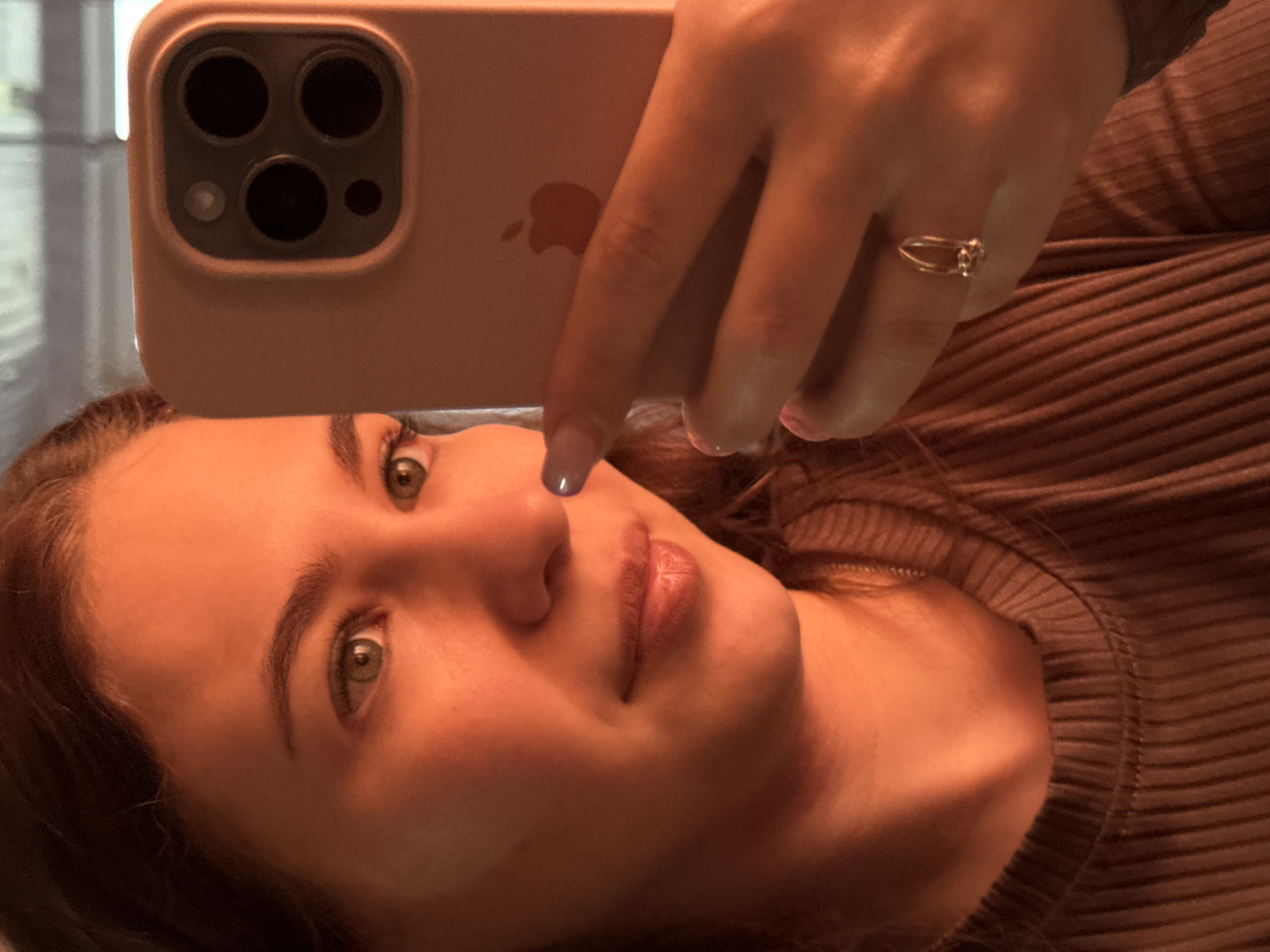 A woman is taking a selfie with a smartphone. The phone has a triple-lens camera and is housed in a light pink case. She is wearing a ring on her finger and a brown ribbed top. The background appears to show a window with blinds. The location is not determinable from the image.
