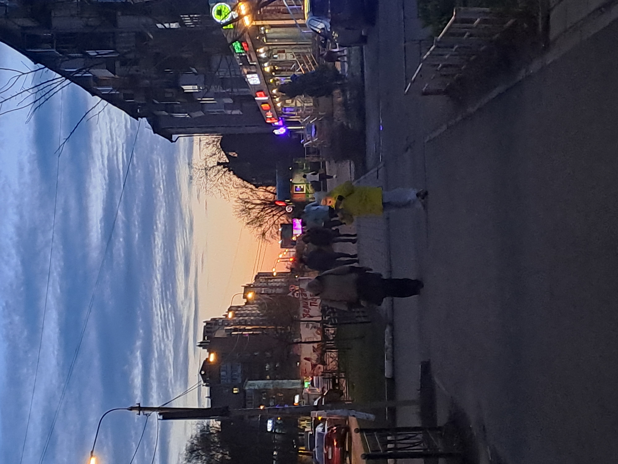 The image depicts a street scene in Elektrostal, Russia at dusk. The sky is a mix of blues and oranges. On the right side, a sidewalk and road are visible. A group of people is walking along the sidewalk, including one person wearing a yellow jacket. On the left, buildings with illuminated signs and windows are present. Power lines stretch across the sky. Several cars are parked along the street. A metal fence or railing is in the lower right corner.