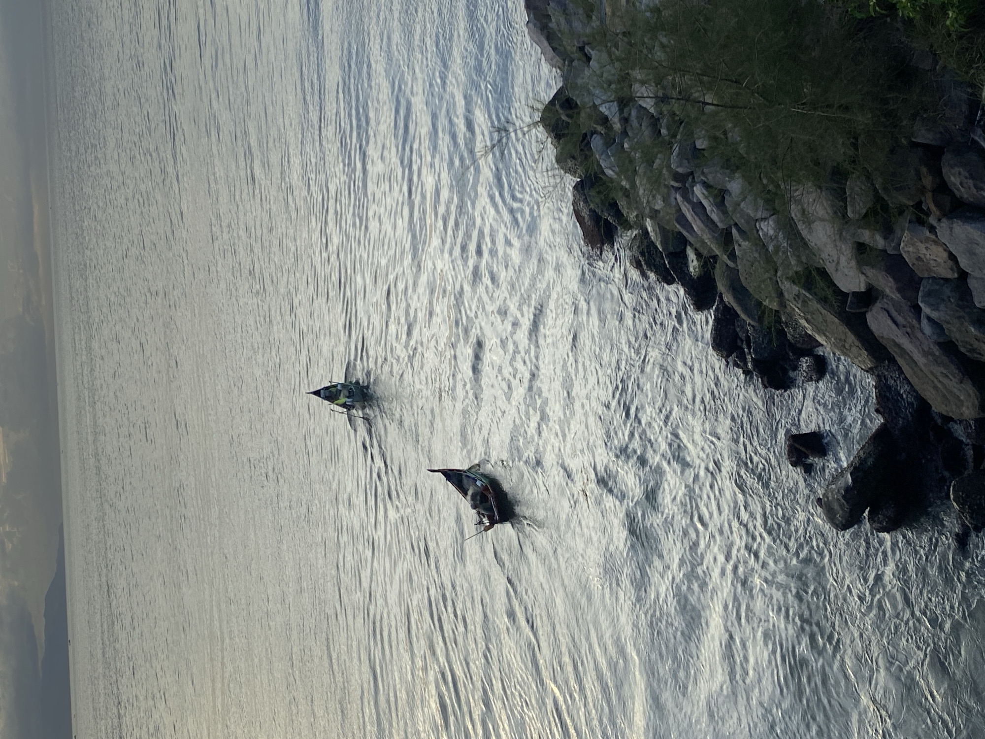 Two small boats are navigating a calm, slightly rippled body of water. The boat in the foreground is dark-hulled with a pointed stern and appears to have one occupant paddling. A second, slightly lighter-colored boat is positioned further in the distance, also with at least one occupant visible, paddling. The water reflects the overcast sky, which is a muted gray with darker clouds on the horizon, obscuring a distant landmass, likely mountains. The scene is framed on the right by a rocky shoreline with sparse, dark green vegetation. The overall mood is serene and slightly somber due to the diffused light. This scene could be located along the coast of Banda Aceh, Indonesia, particularly in an area with a rocky shoreline and open water, possibly near a fishing village or a coastal path.