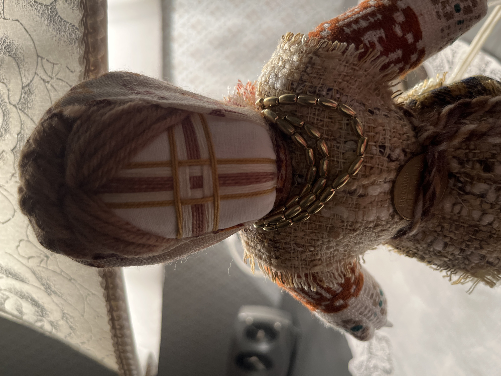 The image shows a close-up, eye-level, portrait-oriented shot of a handmade fabric doll. The doll is positioned centrally in the frame, facing forward, with its head slightly tilted to the right. It is dressed in rustic, textured fabrics. The doll's head is covered with a brown yarn headdress, and its face is a white fabric panel with red and yellow threads forming a cross shape. The doll wears a beaded gold necklace and a belt made of braided string with a coin attached. Its body is covered with a woven fabric vest decorated with orange and white geometric patterns. The background is blurred, but suggests an indoor setting, possibly a kitchen or dining area, with hints of a lamp shade and a kitchen appliance visible. The lighting is warm, coming from the top and right, casting subtle shadows. The doll appears to be sitting or standing on a white surface.