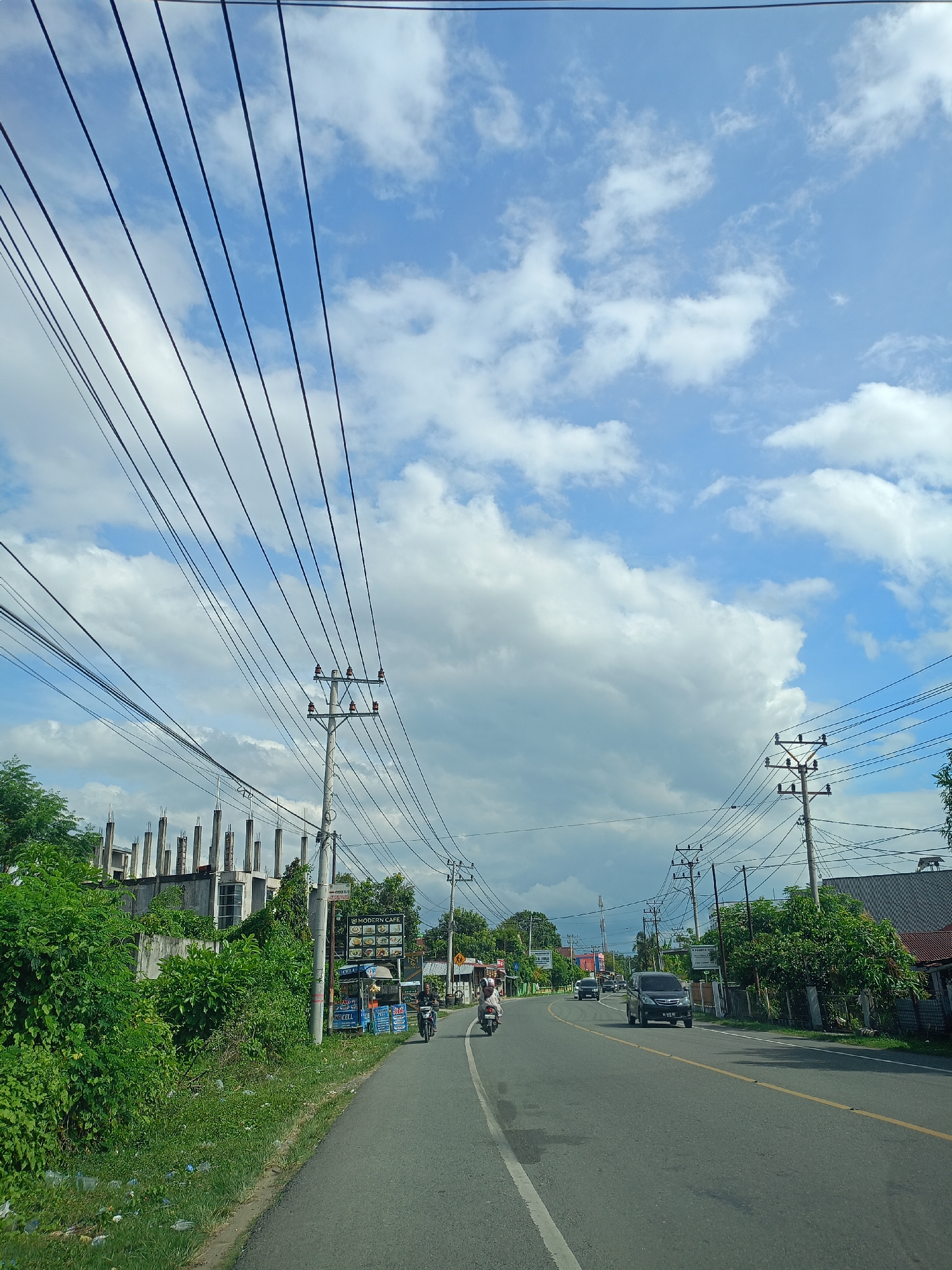 Captured from an elevated perspective, potentially within a moving vehicle, this image presents a street view in Teungoh Baro, Indonesia. The asphalt road, marked by a white lane divider and a yellow side line, leads into the distance, featuring a black SUV and several mopeds. Utility poles, crisscrossed by an array of power lines, line both sides of the road. A building under construction with exposed concrete columns is visible on the left. Lush greenery and vegetation border the road, interspersed with residential houses. A partly cloudy sky occupies the upper half of the frame. A sign for “Modern Cafe” can be seen along the left side of the road.