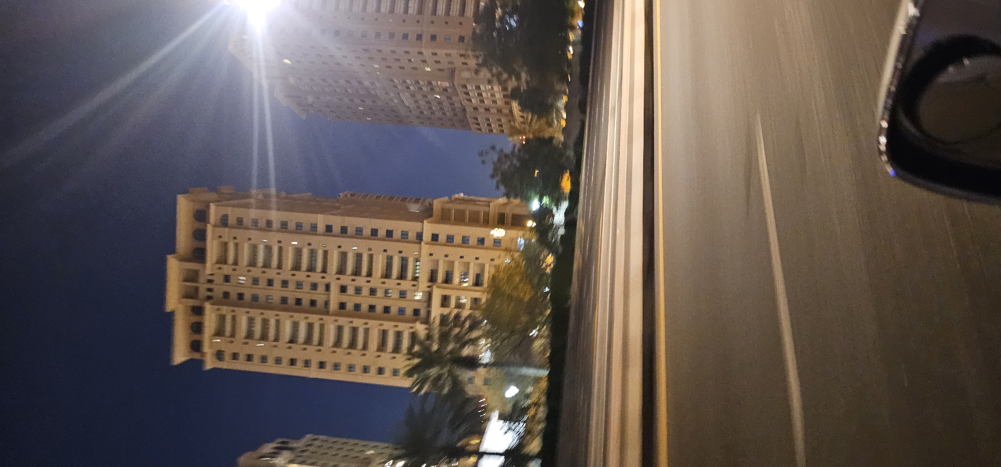 The image shows a nighttime view of high-rise buildings in Doha, Qatar, seen from a moving vehicle. The buildings are illuminated, and the sky is dark blue. In the foreground, a highway with lane markings is visible, suggesting the perspective is from a car driving on the road. Trees and palm trees are also present, indicating a landscaped area alongside the road. The overall impression is of a modern urban environment at night, with the blur from the vehicle's motion adding a dynamic quality to the scene.