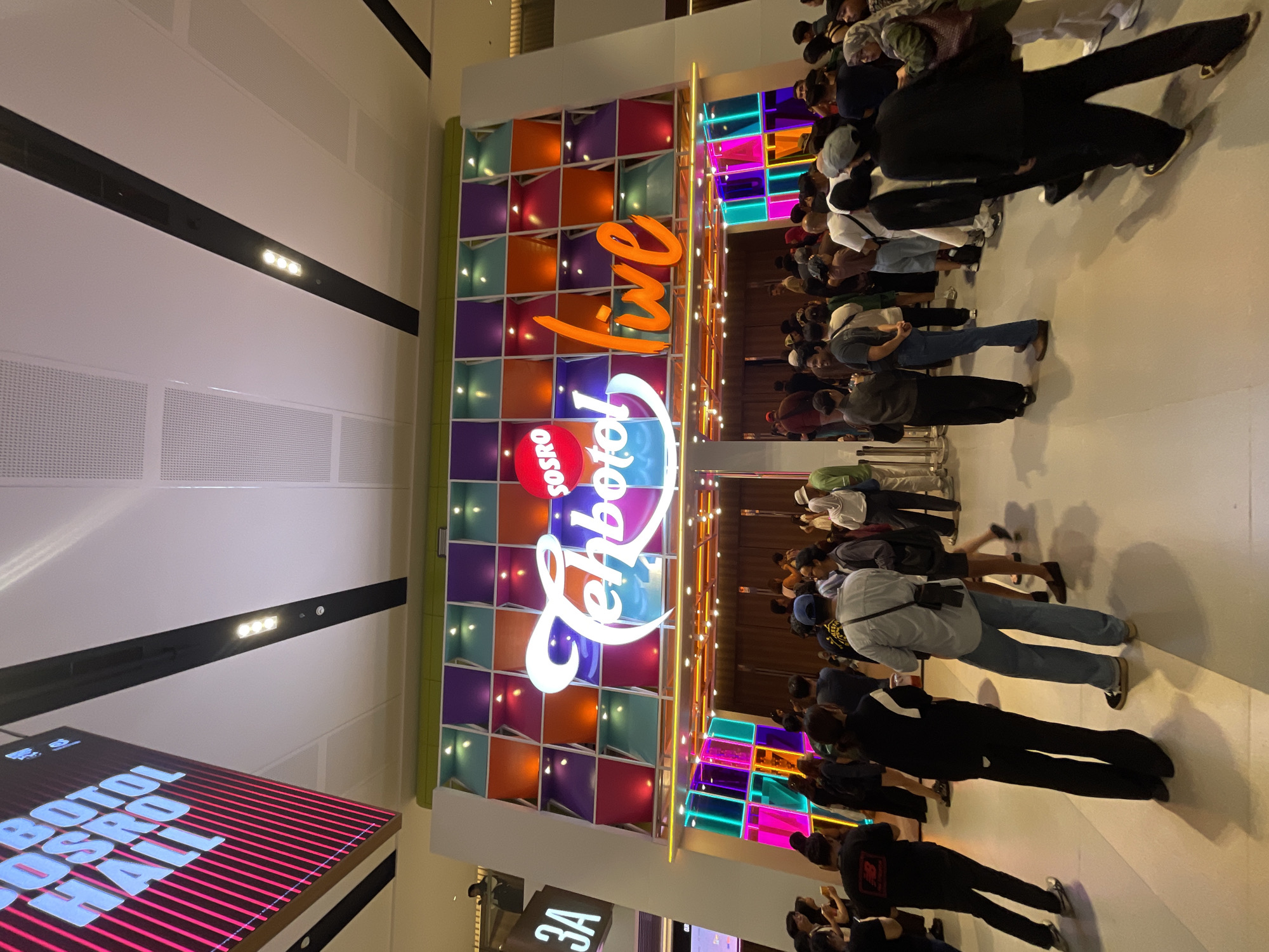 In Salembaran, Indonesia, a queue of individuals extends alongside a wall adorned with a grid pattern of colorful squares. The wall is also illuminated with the phrase "Tehbotol Live" in a stylized design, a reference to the Tehbotol Sosro brand. The crowd appears to be waiting in line, possibly for an event at the venue. The architecture suggests an indoor, commercial space.
