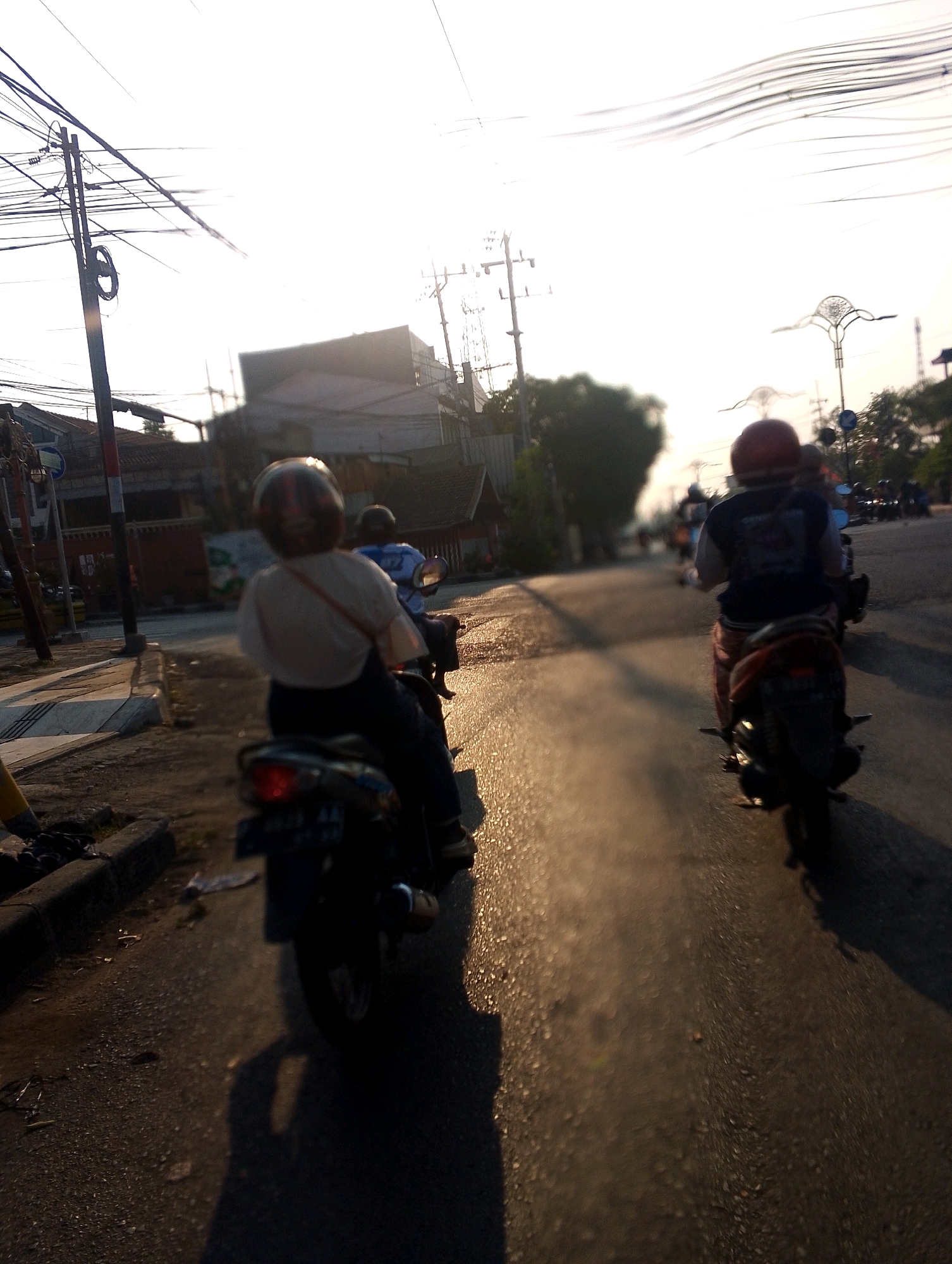 In an outdoor, urban scene under bright sunlight, two people ride motorcycles on a paved road. The foremost motorcycle, on the left, is ridden by a person wearing a red helmet and a light-colored top with dark pants. The motorcycle itself is dark-colored with a license plate visible. The second motorcycle, on the right and slightly further ahead, is ridden by a person wearing a dark shirt with red and white accents and possibly striped pants. This motorcycle also appears to be dark-colored. The road is wet and reflects the sunlight, creating a shimmering effect. Utility poles and wires are visible overhead. In the background, buildings and trees are present. The perspective is from a low angle, likely from another moving vehicle.
