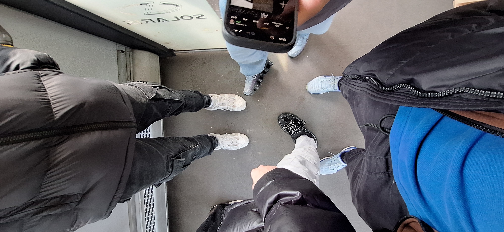 The image shows a group of people standing indoors, with a top-down perspective focused on their feet and lower bodies. They appear to be in a semi-circle.
The individuals are wearing a variety of casual clothing, including puffer jackets and casual pants. The group is taking a photo with a smartphone. The surroundings appear to be a public place, potentially a transit station or enclosed public area. The city mentioned, Burgas, Bulgaria, and the word "Solaris" visible in the upper left suggest the location might be near or inside the "Solaris" mall, a well-known business center in Burgas. The floor is a smooth, neutral-toned material, possibly tile or concrete.