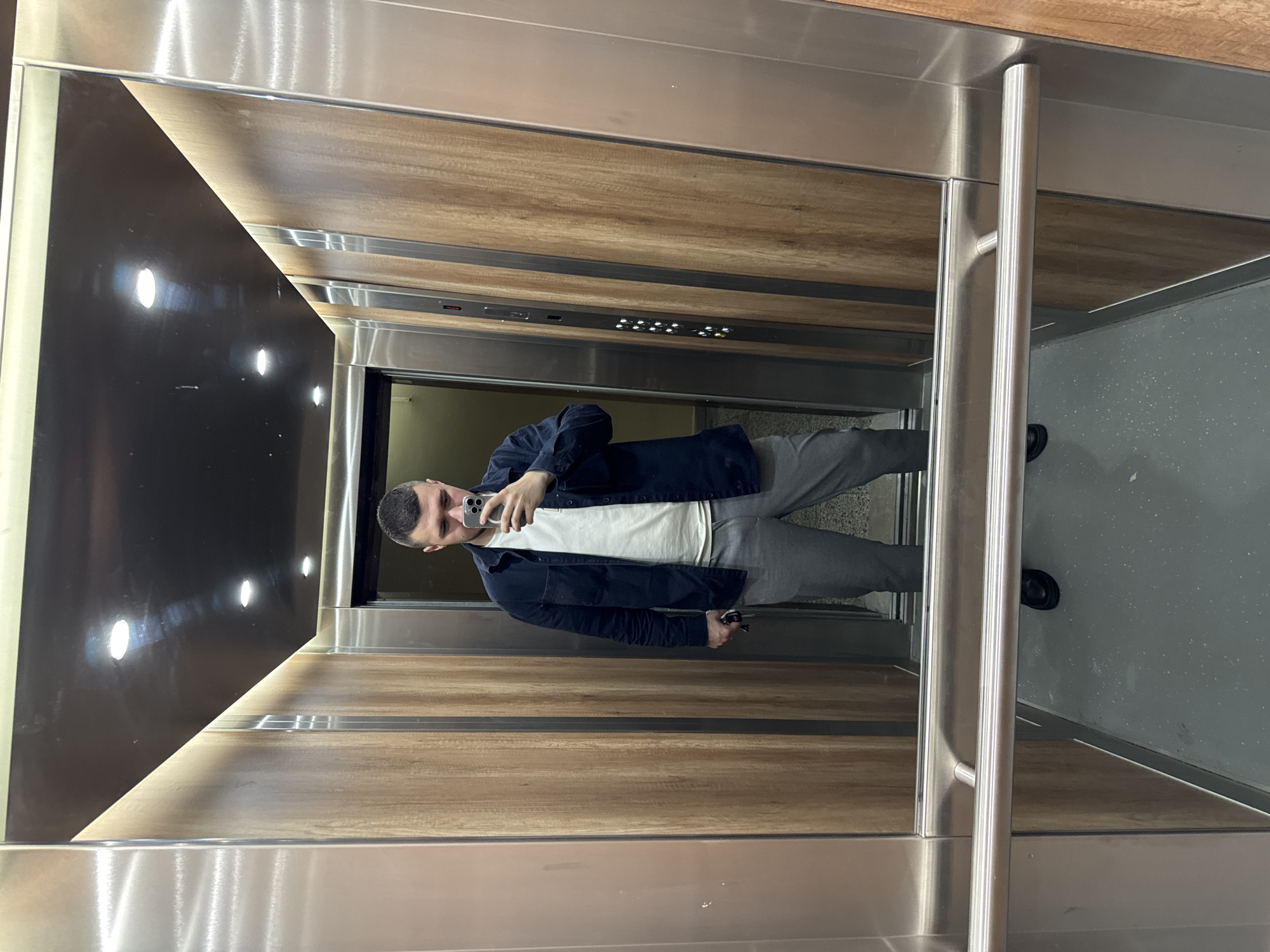 A man takes a selfie in an elevator. He is wearing a white t-shirt, a navy blue button-down shirt, and grey sweatpants. The elevator has wood-paneled walls, a black accent wall with lights, a stainless steel door, and a gray floor. The man is holding his keys in his left hand and his phone in his right hand. The specific location in Sofia, Bulgaria is not discernible from the image.