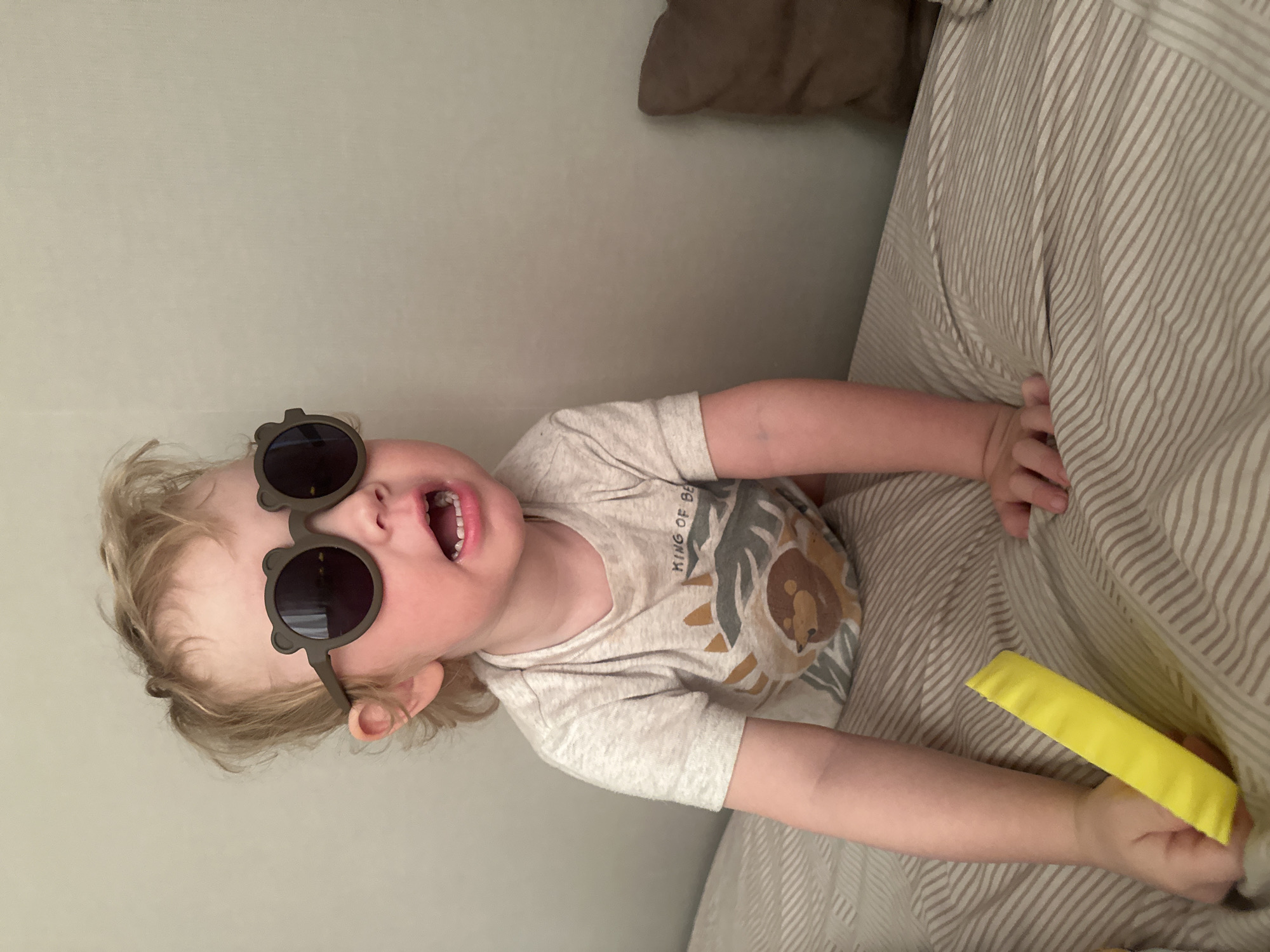 A toddler wearing bear-shaped sunglasses and a shirt with a lion design is lying on a striped bed. The child's mouth is open. The child is holding a yellow object in their hand. The background consists of a plain wall and a brown pillow. There is no information to indicate that this image was taken in Votkinsk, Russia.