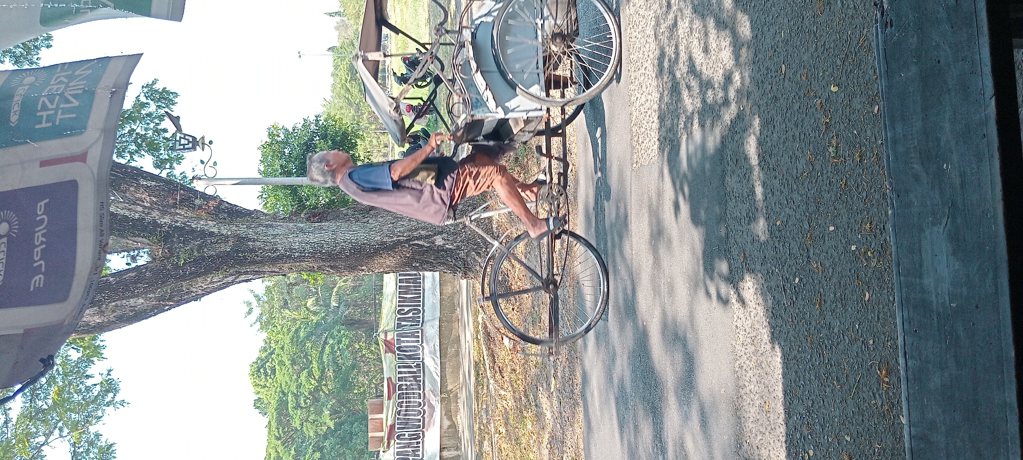 An elderly man is riding a pedicab through a city street. He is wearing a blue t-shirt and brown shorts and has a gray vest over his t-shirt. The pedicab is gray with a canvas top. The man is pedaling and appears to be moving forward. The background shows a large tree, a banner with the text "PANG WOODBALL KOTA TASIKMALAYA", and a grassy area. The sky is bright and clear. The scene appears to be in Tasikmalaya, Indonesia.