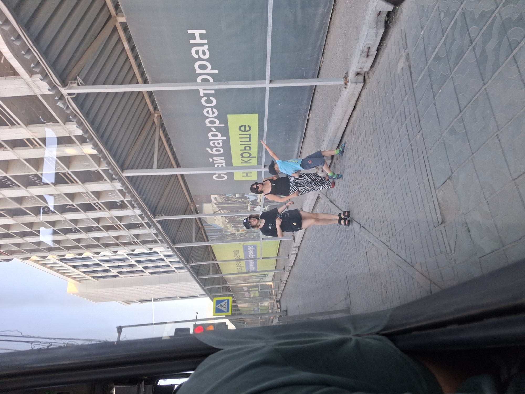 The image depicts an outdoor scene, likely on a sidewalk in Novosibirsk, Russia. In the foreground, a person is partially visible on the left, seen from the shoulder down, seated or leaning against something, with a dark fabric covering their clothing. The surface beneath them appears to be a reflective dark surface, possibly a bus window or a car.
Further into the scene, a paved sidewalk is visible, constructed with grey rectangular paving stones. To the right of the sidewalk, a large, dark grey advertisement banner is stretched across a construction site. The banner features large white Cyrillic text that reads "Сай бар-ресторан" (Sai bar-restaurant) and a smaller yellow rectangular section with Cyrillic text that says "на крыше" (on the roof).
Three individuals are walking on the sidewalk. The first is a young woman wearing a black bucket hat, a black t-shirt with a graphic on it, a black skirt, and black sandals. She is looking towards the camera. Next to her is another woman wearing a black tank top, sunglasses, and a long, patterned skirt with black and white zebra-like stripes, also wearing black sandals.
To the right of the women, a young boy in a blue t-shirt and dark shorts is walking. He is wearing a dark baseball cap and blue sandals. He is actively pointing with his right arm towards the advertisement banner.
In the background, a multi-story building under construction is visible. It has a grid-like facade with many open window frames. A traffic light with red and yellow lights illuminated is present on a pole to the left of the sidewalk, near a blue and yellow pedestrian crossing sign. The overall environment suggests a city street with ongoing construction.