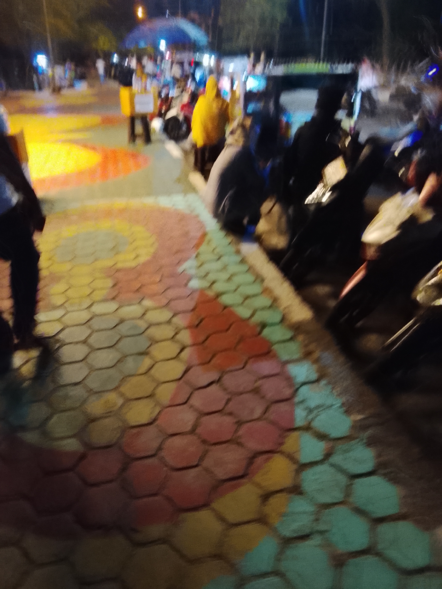 The image shows a blurred scene at night in a public area, likely a marketplace or gathering space, in Tanjung Balai Karimun, Indonesia. The ground is paved with hexagonal tiles painted with colorful patterns, including yellow, orange, red, and teal. Several motorcycles are parked on the right side of the image. In the background, blurry figures of people can be seen, some wearing yellow and others in dark clothing. There are also lights and structures that suggest a lively environment. One person in the foreground is wearing dark pants and a light-colored shirt. Another person, further back, is wearing a bright yellow jacket and holding a cart or stand. The overall atmosphere appears to be one of a busy evening in a local community space.