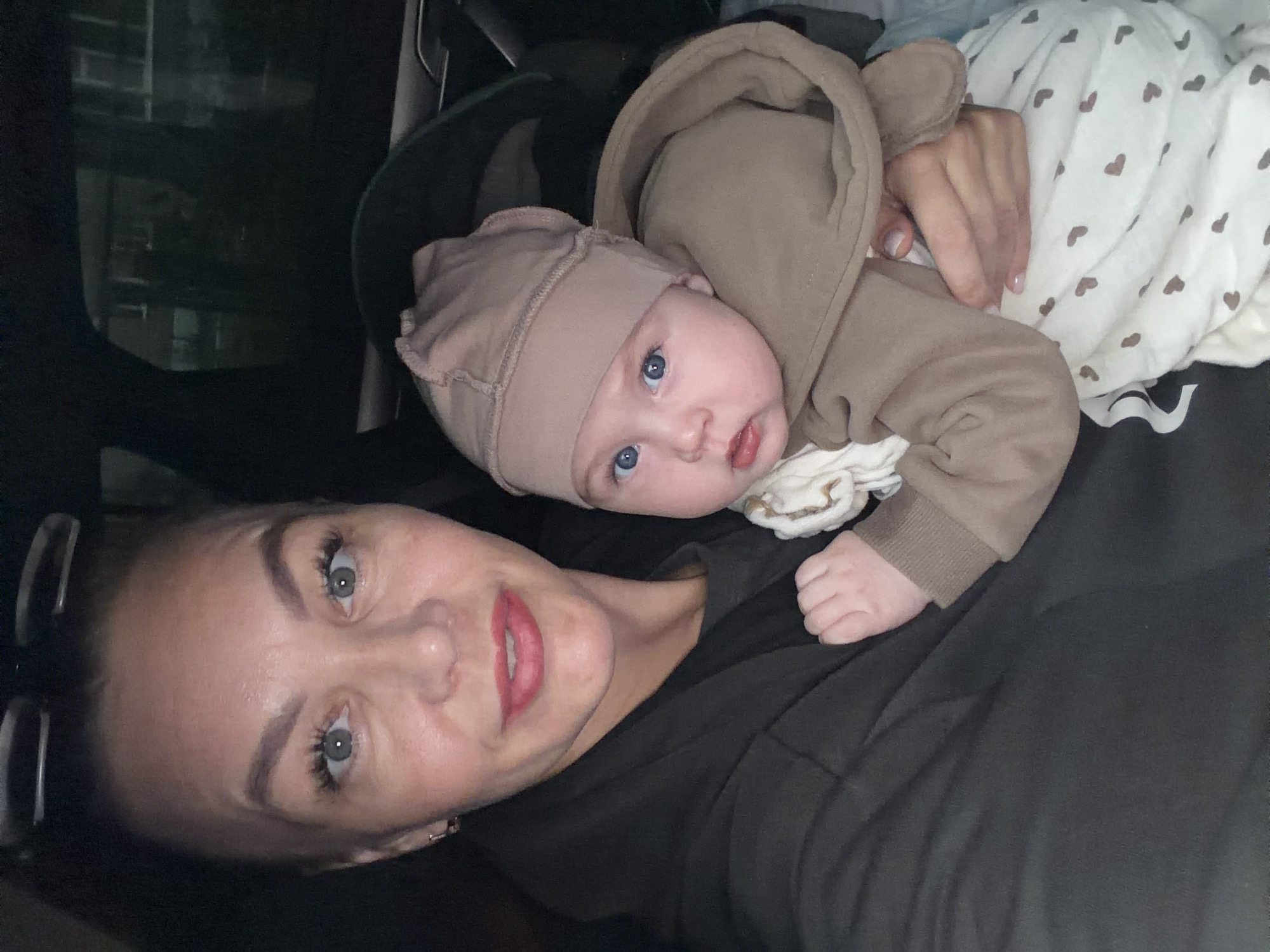 This media file is a close-up, selfie-style photograph featuring two individuals: a woman and a baby. The woman, positioned in the foreground, has light-colored eyes, dark eyebrows, and is wearing subtle makeup, including pink lipstick. Her hair appears dark and is pulled back. The baby, nestled on the woman's chest and shoulder, is wearing a light brown hooded onesie with ear-like details on the hood and a matching cap. The baby has striking blue eyes and a light complexion. The background is dark and appears to be the interior of a vehicle, suggested by the faint reflection of what looks like buildings visible through a window on the left side. The overall lighting is dim, implying it could be nighttime or a dimly lit environment. The baby is wrapped in a white blanket adorned with a repeating pattern of small brown hearts. The emotional tone is intimate and affectionate, with both subjects appearing relaxed and content. No visible text is present in the image. The location context is Chaikovsky, Russia.