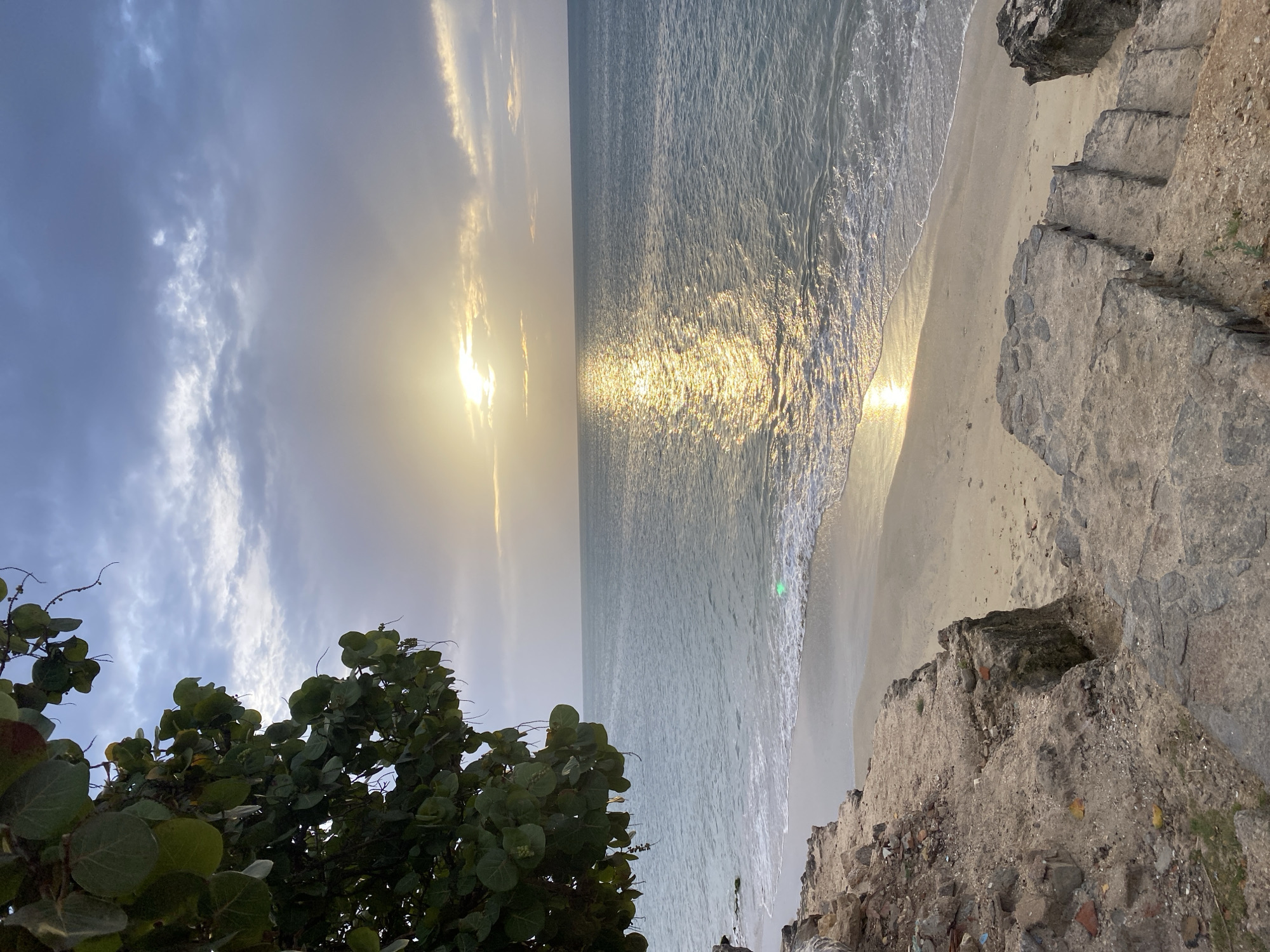 This image depicts a coastal scene at sunset in Juan Griego, Venezuela. The foreground features a weathered stone staircase leading down to a sandy beach. To the left, a rocky embankment is covered in dirt and debris, with a tree bearing large, round green leaves hanging over the scene. The beach is narrow and curves gently along the waterline. The ocean occupies the majority of the frame, its surface reflecting the golden light of the setting sun. The water is a tranquil blue-grey, with small waves gently lapping the shore. The sky above is dramatic, filled with a mixture of dark grey clouds and lighter patches where the sun is breaking through, casting rays of light across the water. The overall impression is one of natural beauty and peacefulness as the day draws to a close.