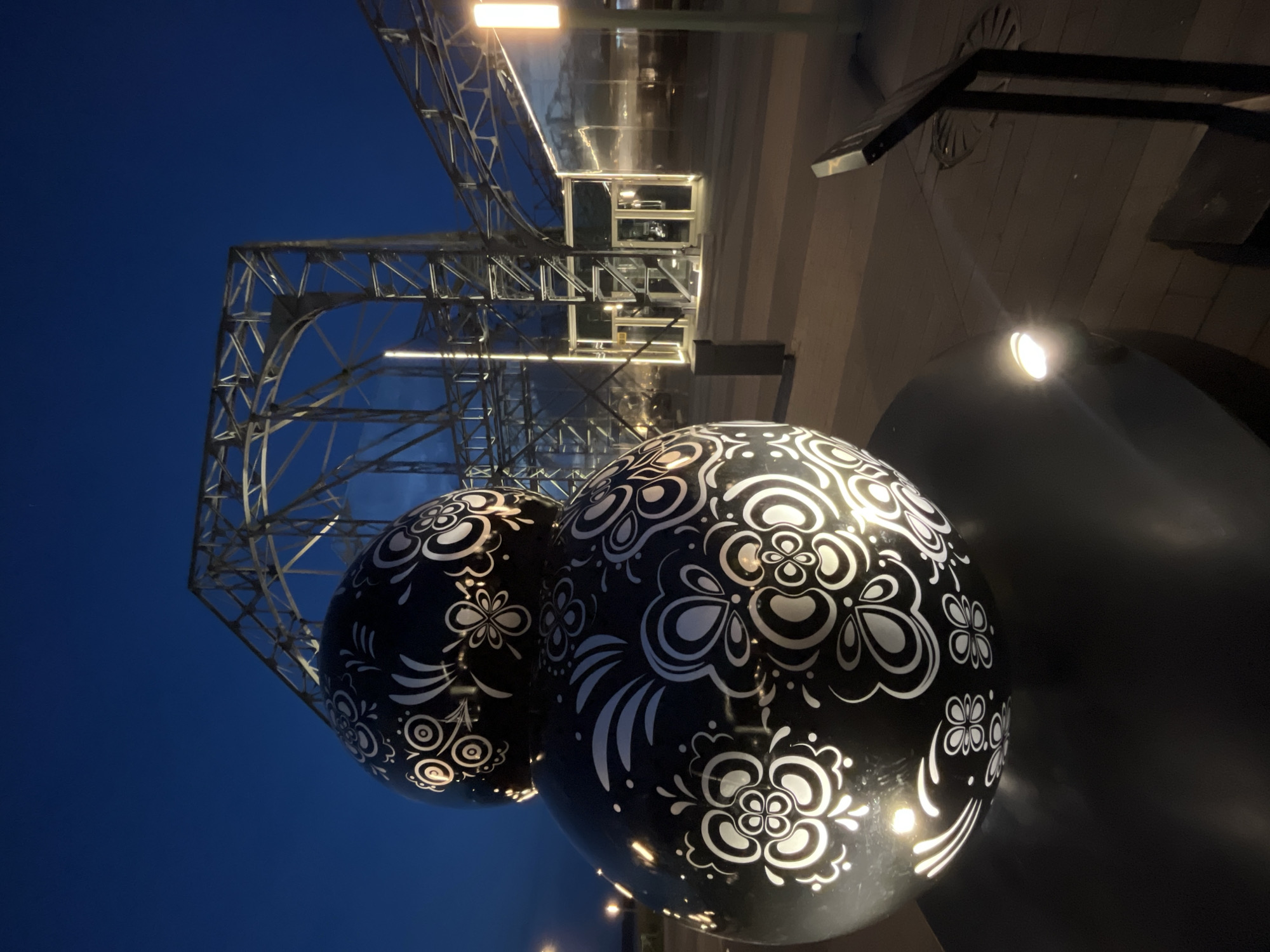 This is a nighttime, low-angle, vertical, full shot of an outdoor art installation in Nizhny Novgorod, Russia. The artwork is a stacked sphere sculpture, black with white floral patterns, lit from within. In the background, a large, skeletal metal structure with glass walls and entrances, illuminated by vertical lights, is visible. The ground is paved with wooden planks, and there is a lamppost to the right. The sky is a deep blue, indicating it is dusk or dawn. The scene appears to be at a public venue or park.