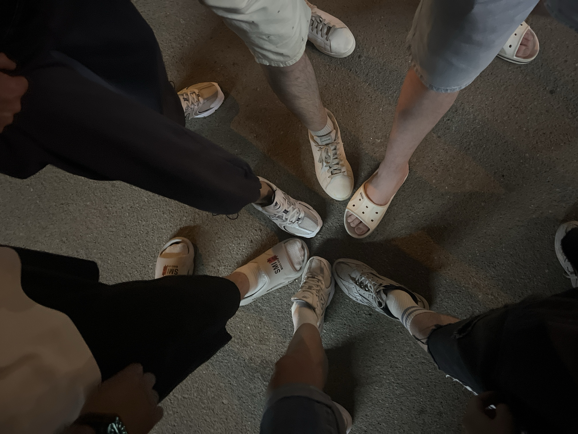 A group of individuals are gathered, their feet and lower legs forming a rough circle on a paved surface. There are 6 individuals. One person is wearing black pants and beige tennis shoes. Another person is wearing dark blue athletic pants and white/grey shoes. One is wearing white/off-white shoes and light grey shorts. One individual is wearing white "crocs". Another person is wearing white tennis shoes, white socks, and blue jeans. One individual is wearing black pants, white socks, and grey athletic shoes. The location is presumed to be in Kazan, Russia.