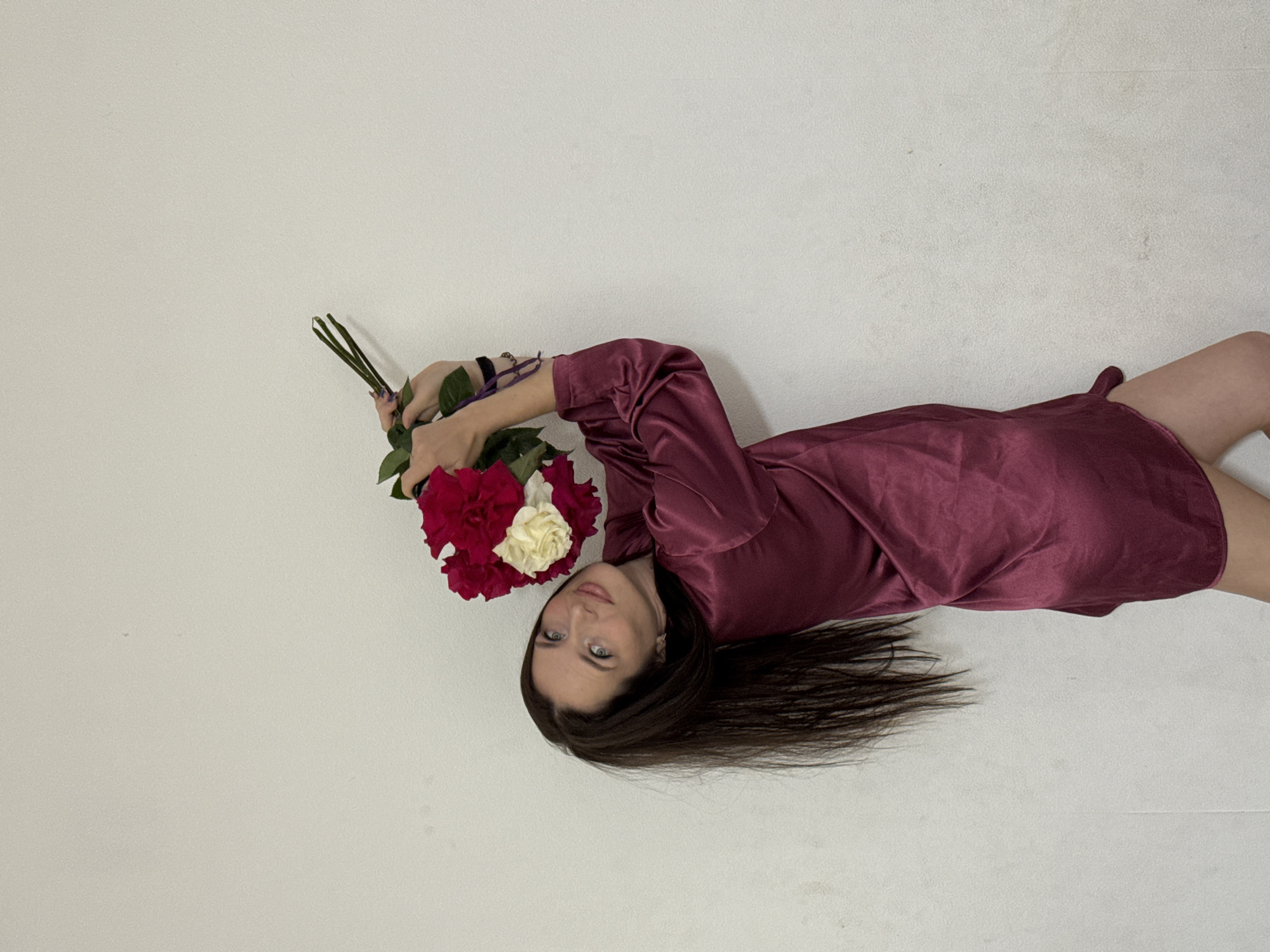 In Rusalevka, Russia, a woman with long dark hair and blue eyes, wearing a burgundy silk dress, stands against a textured white wall. She is holding a bouquet of red and white roses above her head. Her pose is dynamic, with her body tilted and her head turned to look at the camera. The bouquet is tied with a purple ribbon. The lighting is even, suggesting an indoor setting, and the overall atmosphere is serene.