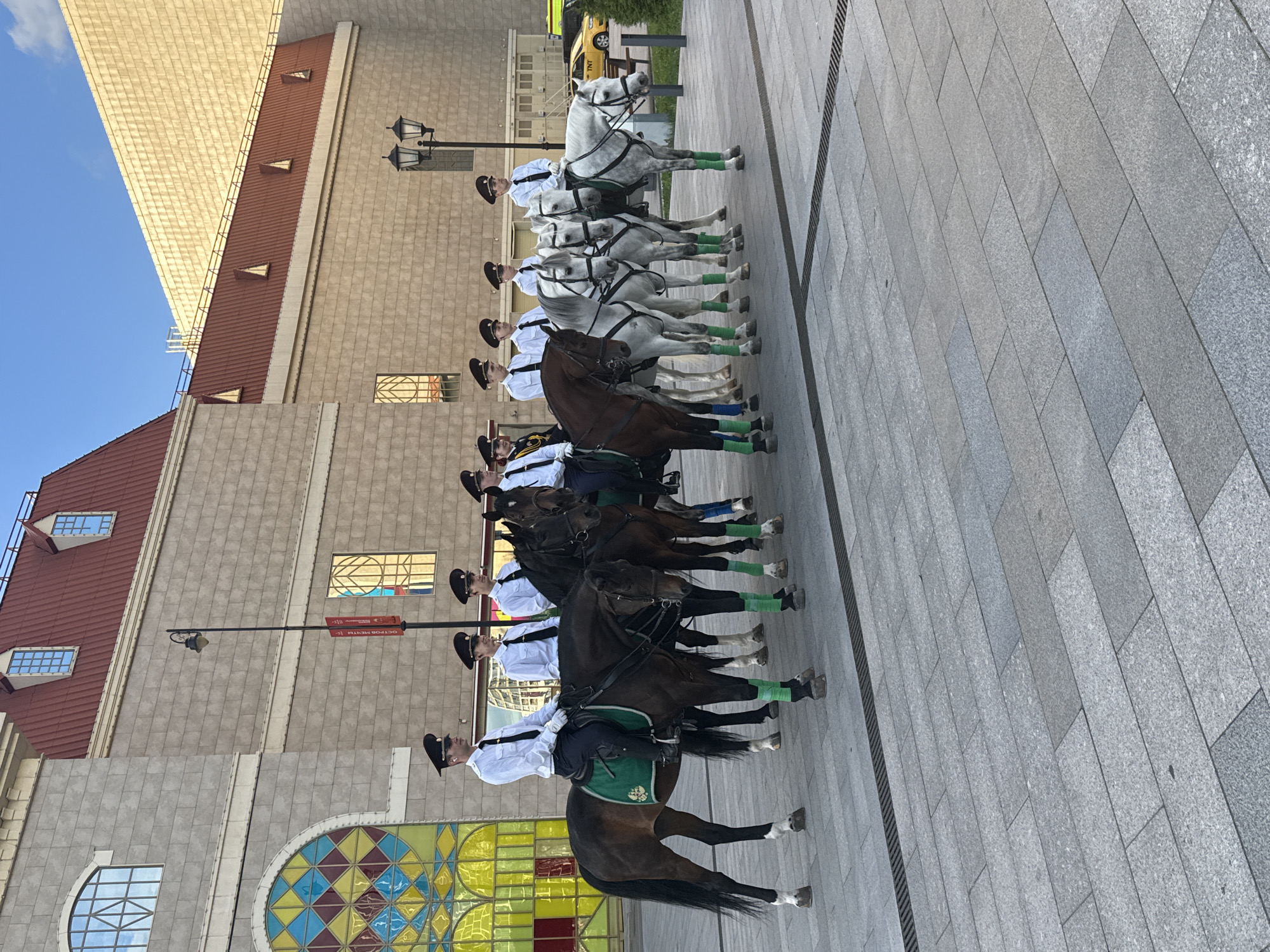 This media file features a group of horse riders, likely a ceremonial or parade unit, positioned in a line on a paved walkway. The riders are all dressed in matching uniforms consisting of white shirts, dark trousers, and dark caps. Many of them are also wearing green leg wraps on their horses. The horses are a mix of white and dark brown. The scene is set outdoors, with a building that has a red tiled roof and a beige tiled facade in the background. There are also architectural details like stained-glass windows visible on the building. The time of day appears to be daytime, with clear skies suggested by the presence of sunlight and shadows. The overall atmosphere seems orderly and formal, consistent with a planned event. Some of the horses have green saddle blankets. The paved walkway appears to be made of large stone tiles. The image is oriented vertically. One visible sign on a lamppost appears to have Cyrillic text, indicating a Russian location.