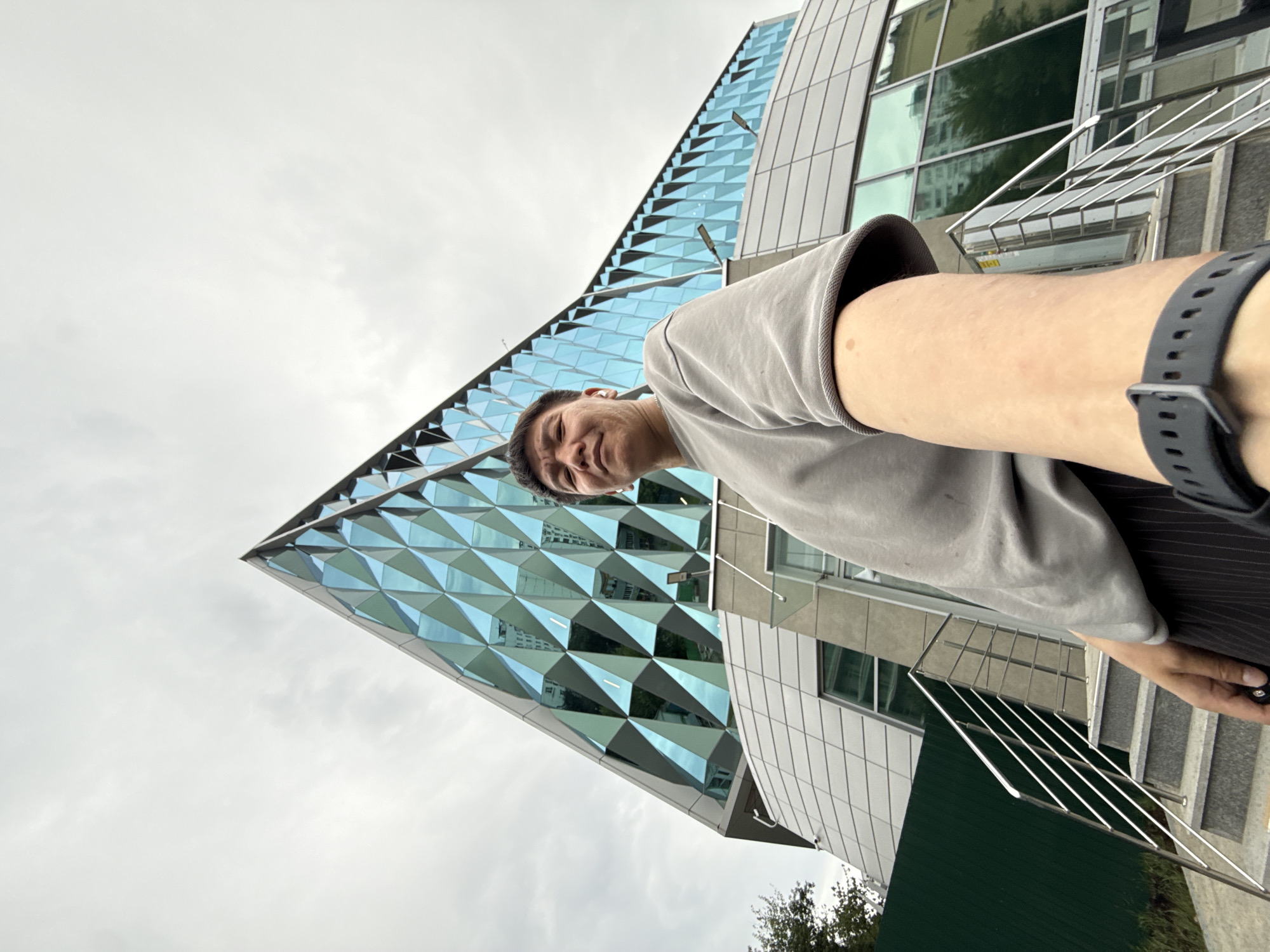 A person is taking a selfie outdoors in front of a modern building. The building features a distinctive facade of geometric, diamond-shaped glass panels that reflect the sky. A staircase with a metal railing is visible to the right of the person, leading up to the building entrance. The person is wearing a grey t-shirt and a black smartwatch. They are smiling at the camera. The sky is overcast, suggesting it might be a cloudy day. The scene appears to be in an urban environment, likely in Moscow, Russia, given the provided context. No visible text is present in the image.