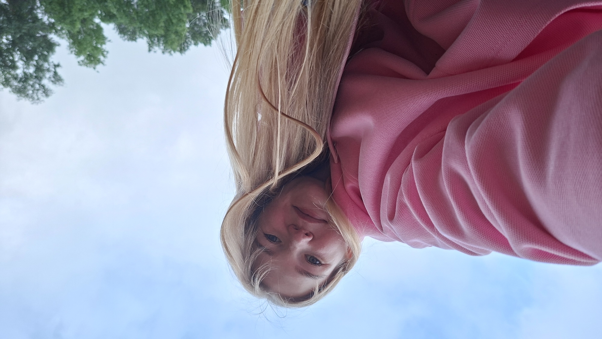 The image depicts a close-up, low-angle perspective of a young individual, likely female, situated outdoors in the city of Chaikovsky, Russia. The subject has long, light blonde hair and is wearing a pink hooded sweatshirt with a textured fabric. Their face is visible, with eyes looking towards the camera, and a subtle, closed-mouth expression. The upper half of the image features a pale blue sky with diffused white clouds. The lower right background reveals a cluster of green deciduous trees, indicating a natural or landscaped environment. The composition suggests the subject is looking down at the camera, potentially taking a selfie.