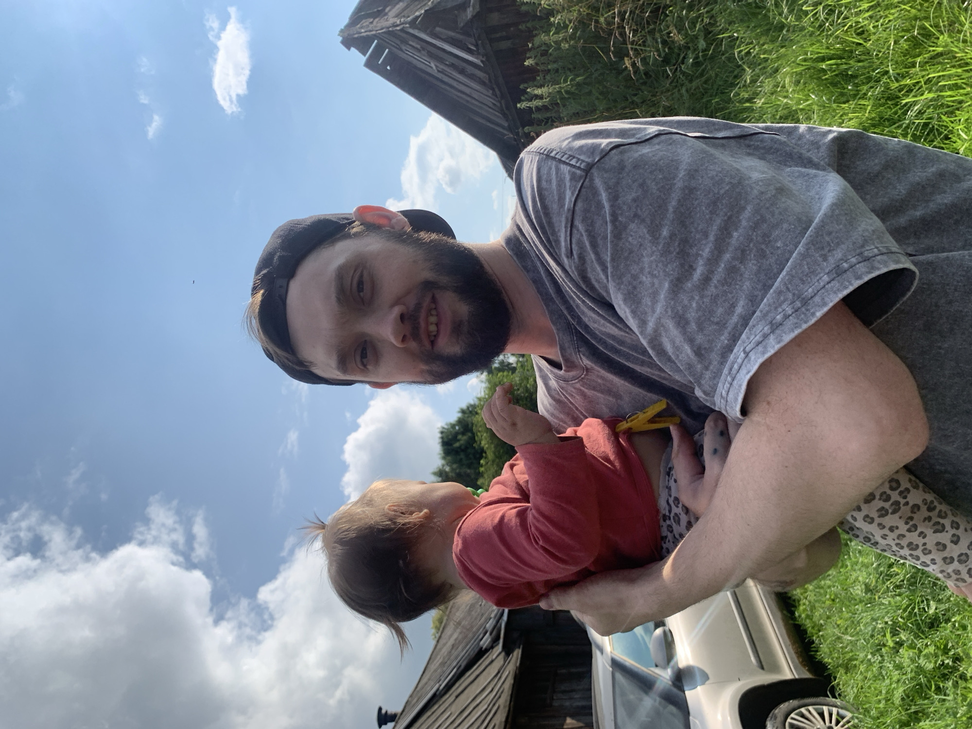 A man wearing a grey t-shirt and a black baseball cap is holding a baby. The baby is wearing a pink long-sleeved shirt and leopard-print pants. The man and baby are standing outdoors, with a car and two wooden structures in the background. The sky is blue with some clouds. The location appears to be a rural area in Zasechnyi, Russia.