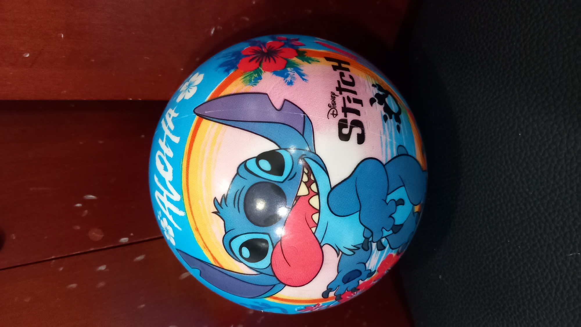 A close-up, eye-level, medium shot captures a Stitch-themed ball resting on a dark, textured surface, likely a seat or table. The ball, predominantly blue and white, features a vibrant illustration of the character Stitch from Disney's Lilo & Stitch. Stitch is depicted with his mouth open, tongue out, and large ears spread. The ball also displays "Aloha" in stylized white lettering across a rainbow gradient background, along with images of tropical flowers and foliage. A "Disney Stitch" logo is visible on the lower right side of the ball. The background is a dark, reddish-brown, possibly wood paneling, with some faint reflections visible. The lighting is natural and direct, casting subtle shadows and highlighting the textures of the ball and the surface it rests on. This image does not depict any elements of the city of Kos, Greece.