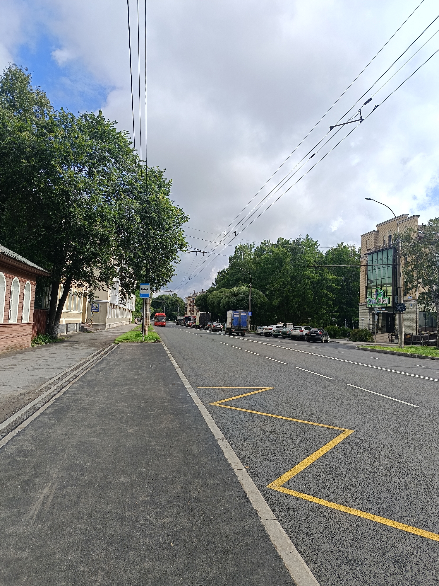 The image shows a street scene in Semenkovo, Russia. A wide asphalt road with multiple lanes is visible, marked with white lane lines and yellow zigzag markings for a bus stop. On the left side of the road, there is a sidewalk with a paved path and a grass verge. Adjacent to the sidewalk is a low wooden building with arched windows, painted red. Further down the sidewalk, a bus stop sign is present next to a red bus. Behind the bus stop and sidewalk, there are several multi-story buildings and a densely wooded area with tall trees. Overhead, trolleybus wires are suspended across the street, supported by poles. Several cars and a large truck are parked along the side of the road. The sky is cloudy with patches of blue sky visible. The overall scene depicts a typical urban street in a town or city, with elements of residential buildings, commercial establishments, public transportation infrastructure, and natural greenery.