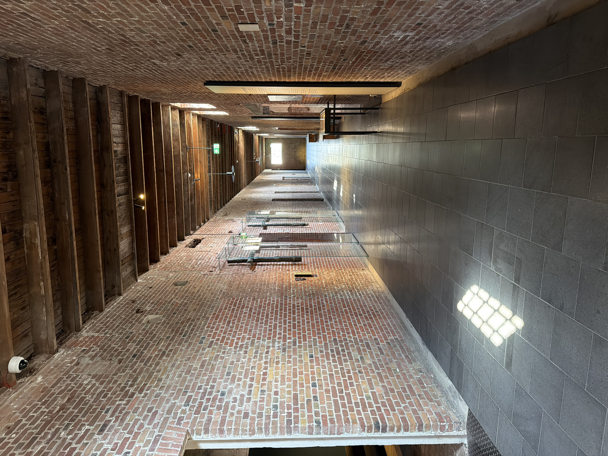 The image shows a long, narrow brick hallway in what appears to be a museum or historical building. The walls are constructed of red brick, and the ceiling consists of exposed wooden beams. The floor is tiled with large, dark grey square tiles. Sunlight streams in from windows along the right side of the hallway, casting a diamond-shaped pattern on the floor. Along the walls are various exhibits, including what appear to be framed pictures or informational displays. On the left side, there are doorways leading into other rooms. A security camera is visible on the ceiling in the upper left corner. A green exit sign with an arrow is illuminated overhead further down the hallway. The environment is sterile and well-lit, suggesting a public indoor space. This hallway is located in the **National Monument Camp Vught**, situated in the municipality of Vught, North Brabant, Netherlands. It is not located in The Hague.