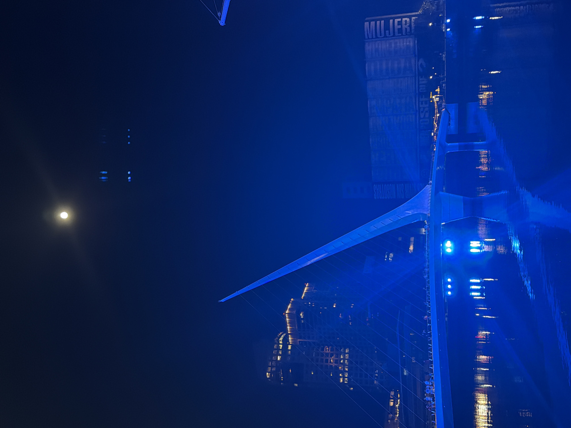 The image depicts the Puente de la Mujer (Woman's Bridge) in Buenos Aires, Argentina, at night. The bridge, illuminated with blue light, is reflected in the water below. A full moon is visible in the dark sky. A building with the word "MUJER" (woman) is situated next to the bridge.