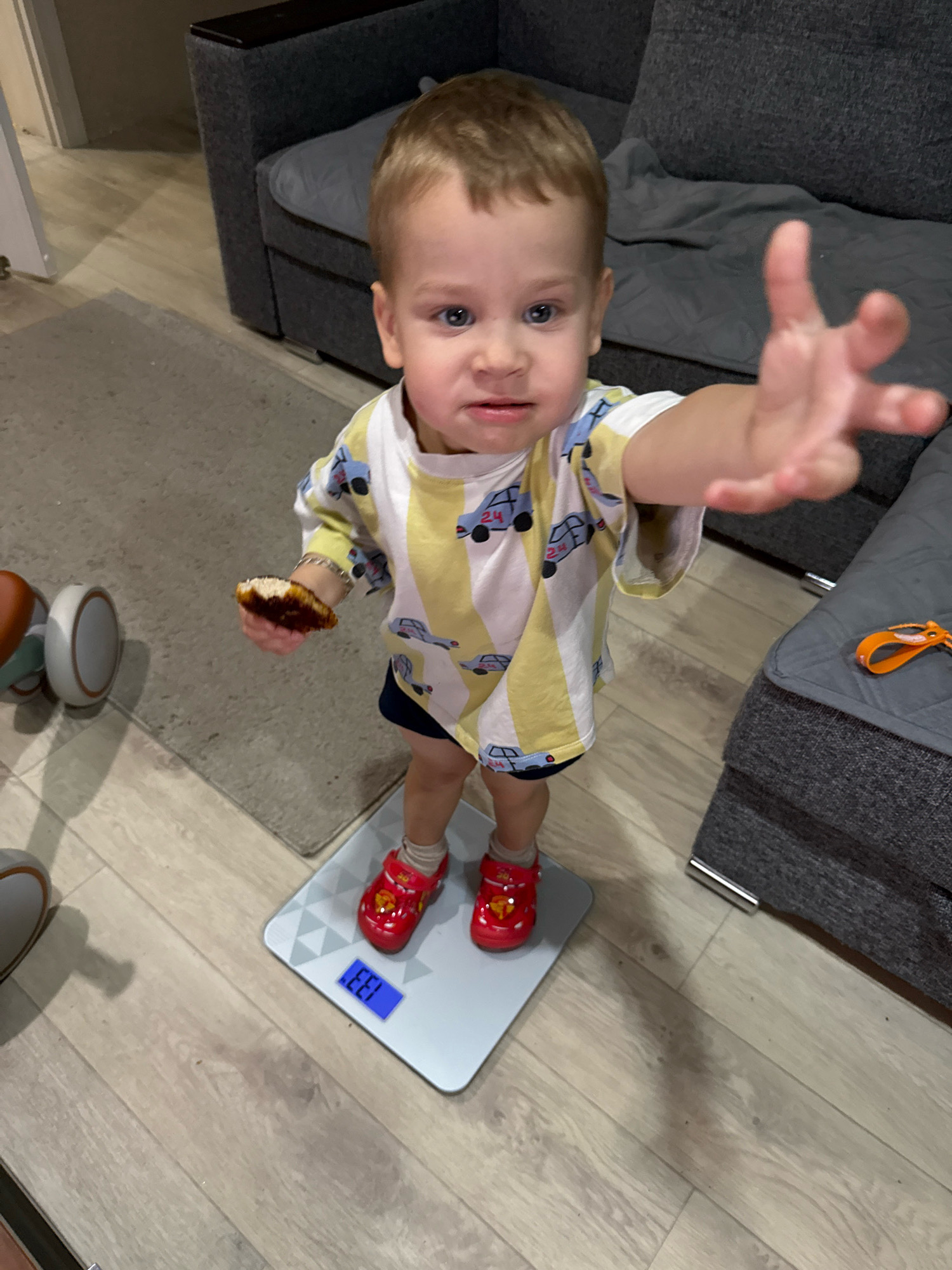 A young child with blonde hair and blue eyes is standing on a digital scale in an indoor setting. The child is wearing a yellow and white striped shirt with car graphics and red shoes. The child is holding a piece of food in their left hand and reaching out with their right hand. The scale displays "13.3". In the background, a grey sofa and a rug are visible. The flooring appears to be wood laminate. There is no visible indication of the city of Rusanovo, Russia.