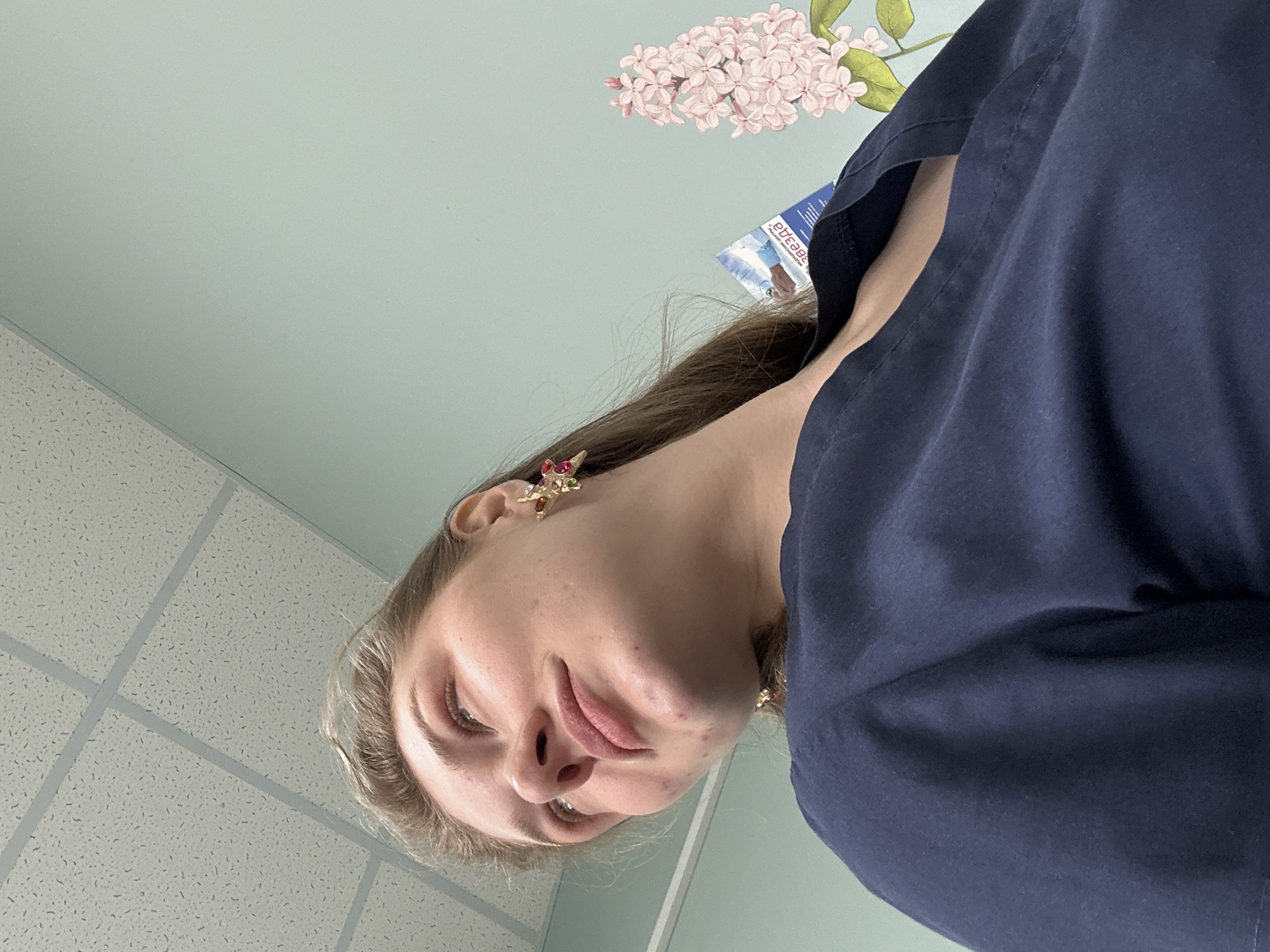 A low-angle, tilted selfie of a person with fair skin and light brown hair. The person is wearing a dark blue garment and has an ornate gold earring. The background consists of a pale green wall adorned with a flower decal. Sections of a suspended ceiling are visible in the lower-left. A piece of paper is seen near the person's neck. The location in Kazan, Russia, cannot be determined from the visual information provided.