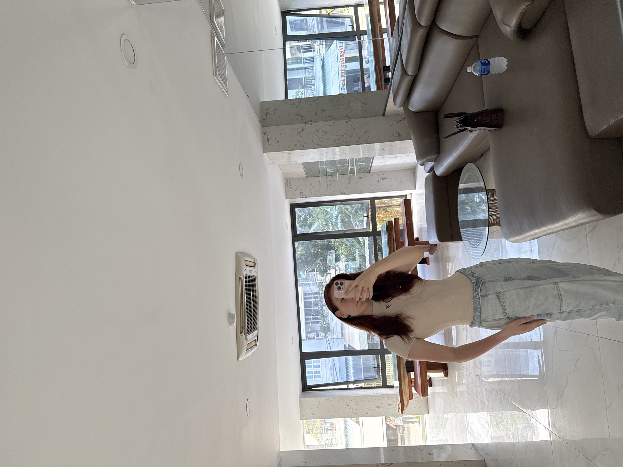 A woman with long red hair is taking a selfie in a modernly decorated apartment in Bac Nha Trang Ward, Vietnam. She is wearing a beige top and light blue jeans. She is holding a pink smartphone to take the picture. The room has large windows overlooking an urban area, a cream-colored leather sofa, a glass-topped coffee table, and an air conditioning unit installed high on the wall. The room has minimalist decor with a white wall and ceiling. A bottle of water and a brown clutch are on the sofa. The floor is light-colored marble.