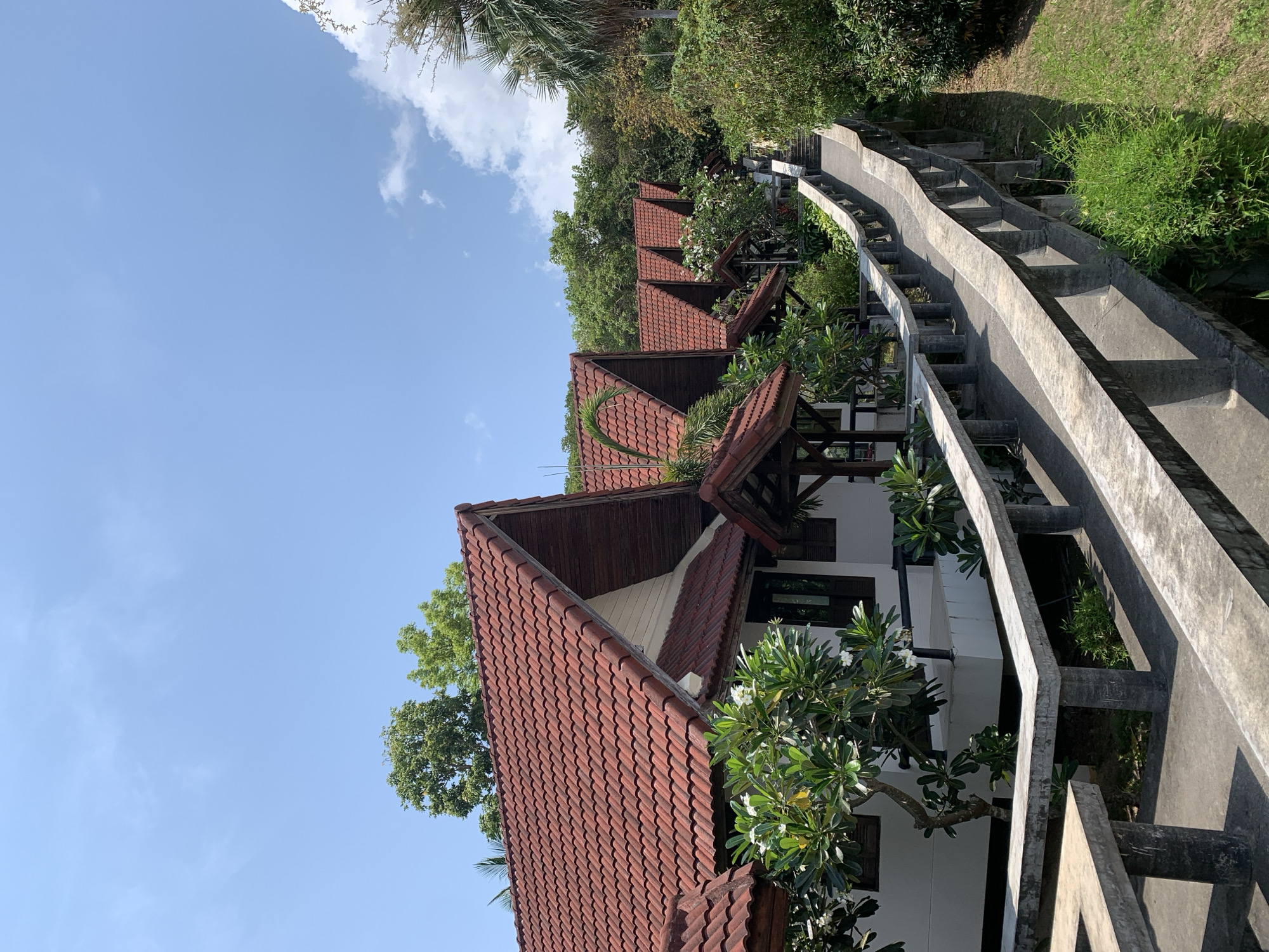 The image captures a serene hillside scene, likely a resort or residential area, in บ้านบางรักษ์, Thailand, viewed under clear daylight. Several charming bungalows are arranged along an ascending slope, each featuring a distinctive red-orange tiled roof and white or light-colored walls. Their design includes traditional dark wooden eaves and window frames, suggesting local architectural influences.
Lush tropical vegetation, including dense green trees, bushes, and vibrant flowering plants, surrounds and partially obscures the bungalows. Some of these plants, possibly plumeria, bear white blossoms, adding to the picturesque setting.
A prominent feature is a long, winding concrete pathway or staircase that ascends the hillside alongside the buildings. It is equipped with a sturdy concrete railing on both sides, which has a unique segmented or wavy design. Strong, distinct shadows are cast on the steps and railing, indicating bright, direct sunlight, suggesting the time of day is either mid-morning or early afternoon.
Above the landscape, a clear, expansive blue sky is visible, adorned with only a few wispy white clouds. The overall atmosphere conveys tranquility and the beauty of a tropical environment. No people or specific events are depicted, and there is no visible text within the frame.