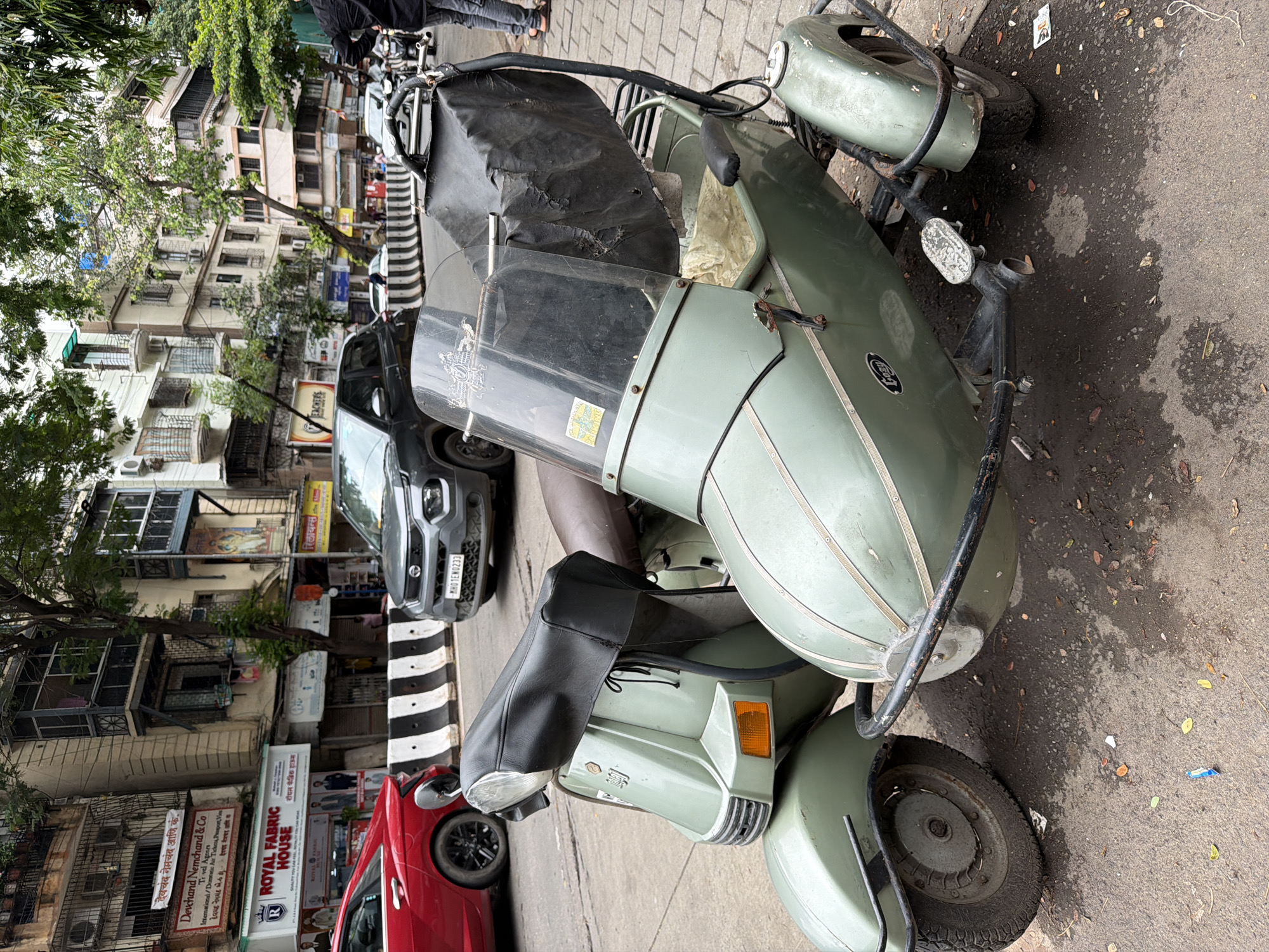 A light green scooter with a sidecar is parked on a city street. The scooter has a black seat cover and a black tarp over the sidecar. The sidecar is also light green and has a black metal frame. The street is paved with asphalt and there are some leaves and debris on the ground. In the background, there are several buildings, some of which appear to be shops. A red car is parked to the left of the scooter, and a gray SUV is parked in the background. The scene appears to be in Mumbai, India, given the architecture and the presence of the Tata Motors logo on the SUV.
