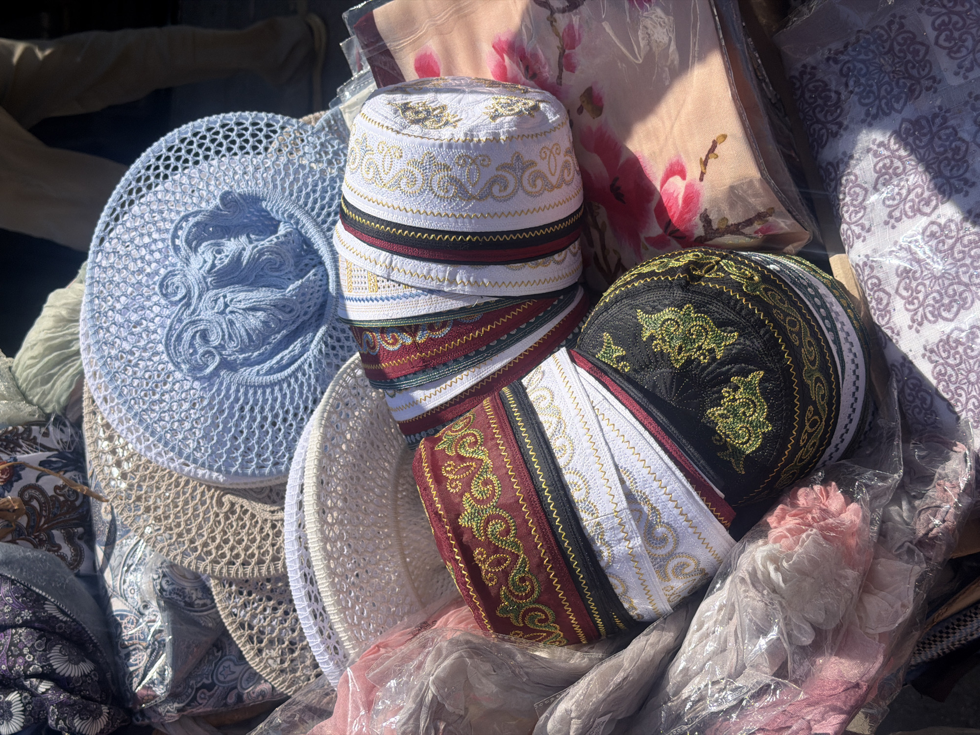 The image displays a close-up view of various hats and fabrics at a market stall, likely in Kanash, Russia. In the foreground, several skullcaps with intricate embroidery in red, white, and black are stacked. To the right of the skullcaps is a light-colored fabric with a floral pattern. In the background, there are woven hats, including a pale blue crocheted hat with an elephant design and a beige crocheted hat. The scene appears to be outdoors, with natural light illuminating the items. The presence of market stalls and the variety of textiles suggest a commercial setting.