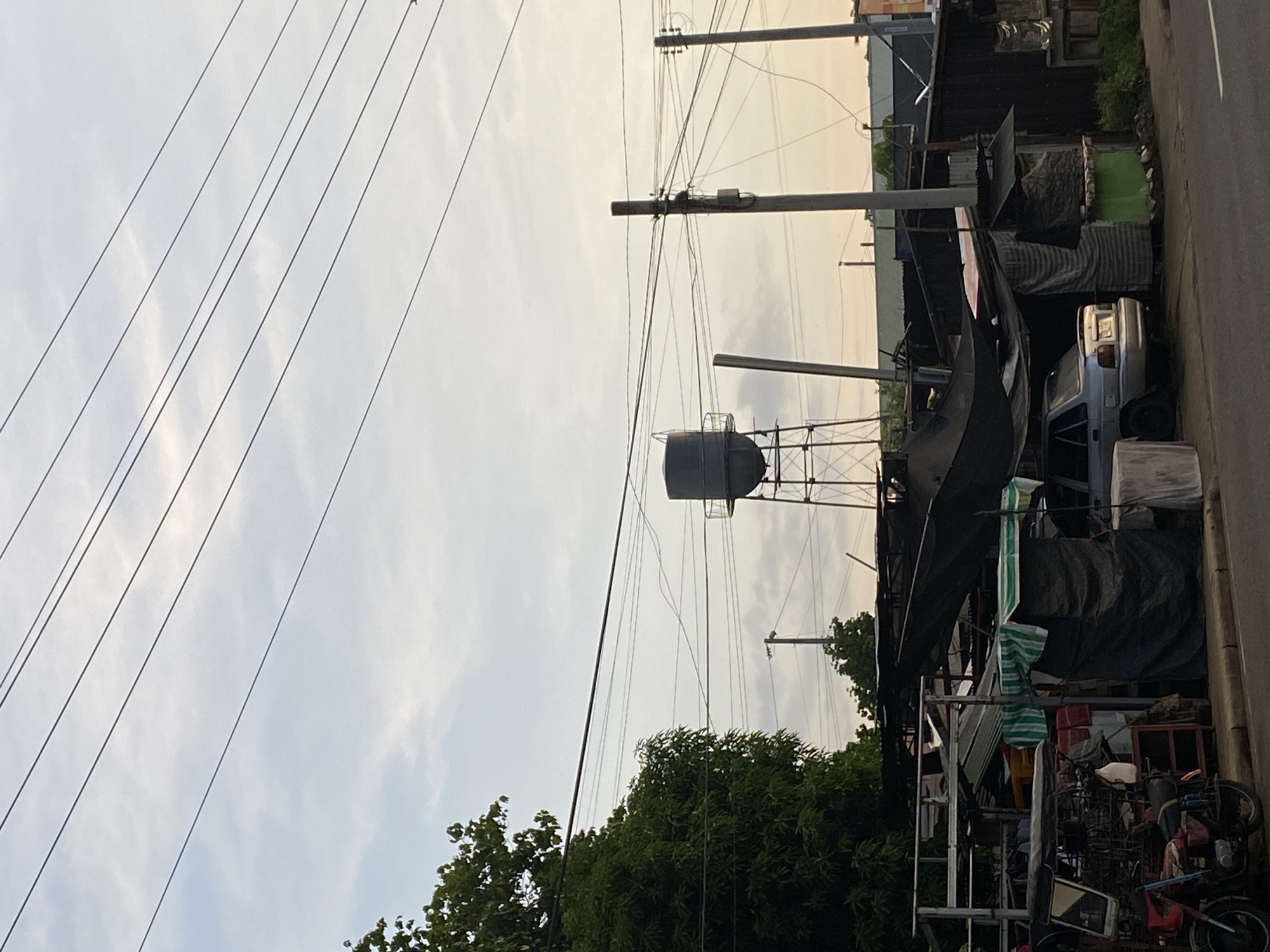 The image depicts a street scene in Davao City, Philippines, likely during late afternoon given the soft, diffused sunlight and the pale sky with scattered clouds. Dominating the background is a tall water tower supported by a metal frame. In the foreground, a roadside scene unfolds with several structures, some appearing to be makeshift stalls or informal dwellings, covered with tarpaulins and corrugated metal. A silver sedan is parked partially under one of these covered areas. To the left, a red tricycle and motorcycle are visible, suggesting local transportation. Overhead, a network of power lines crisscrosses the sky, a common sight in urban and suburban areas. The overall impression is one of everyday life in a bustling, potentially developing area, with a blend of infrastructure and informal settlements.