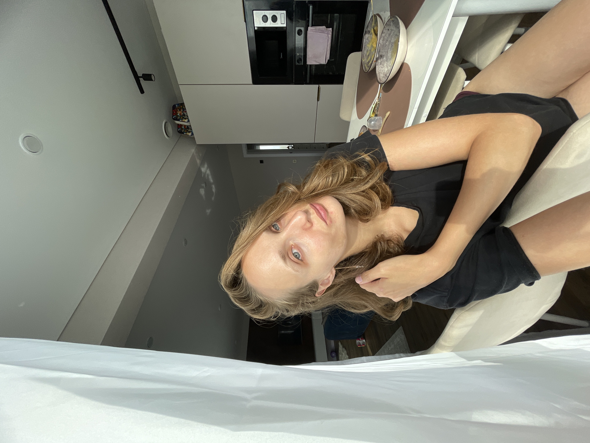 In this media file, a young woman with blonde, wavy hair and blue eyes is positioned in a kitchen setting. She is wearing a black t-shirt and dark shorts. The background reveals modern kitchen cabinetry, a built-in oven with a microwave above it, and a dining table with chairs. Two decorative plates with intricate designs are visible on the table. The scene appears to be indoors, likely during the daytime, as indicated by the natural light filtering in, casting shadows. The woman is looking towards the camera with a calm expression, resting her head on her hand. There is no visible text in the image. The overall impression is a casual, domestic moment captured in a contemporary living space.