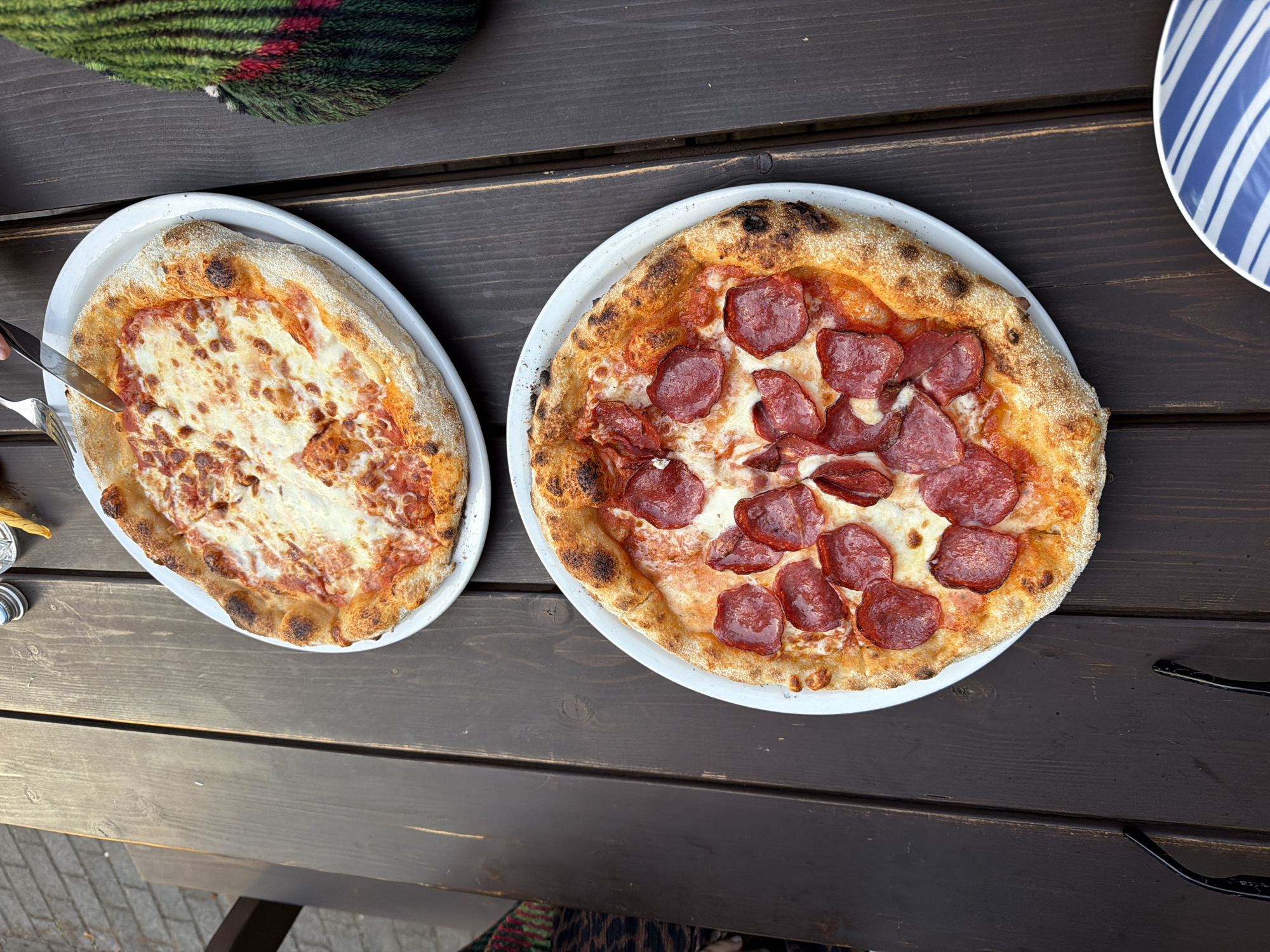 Two pizzas are on a dark wooden table outdoors. The pizza in the foreground is a pepperoni pizza with melted cheese and red pepperoni slices. The pizza in the background appears to be a margherita pizza with tomato sauce and melted cheese. There is a fork and knife cutting into the margherita pizza. A striped plate is visible in the bottom right corner. The scene is set in the city of Ventspils, Latvia, possibly at an outdoor dining establishment near the beach or harbor, given the picnic table and general atmosphere.