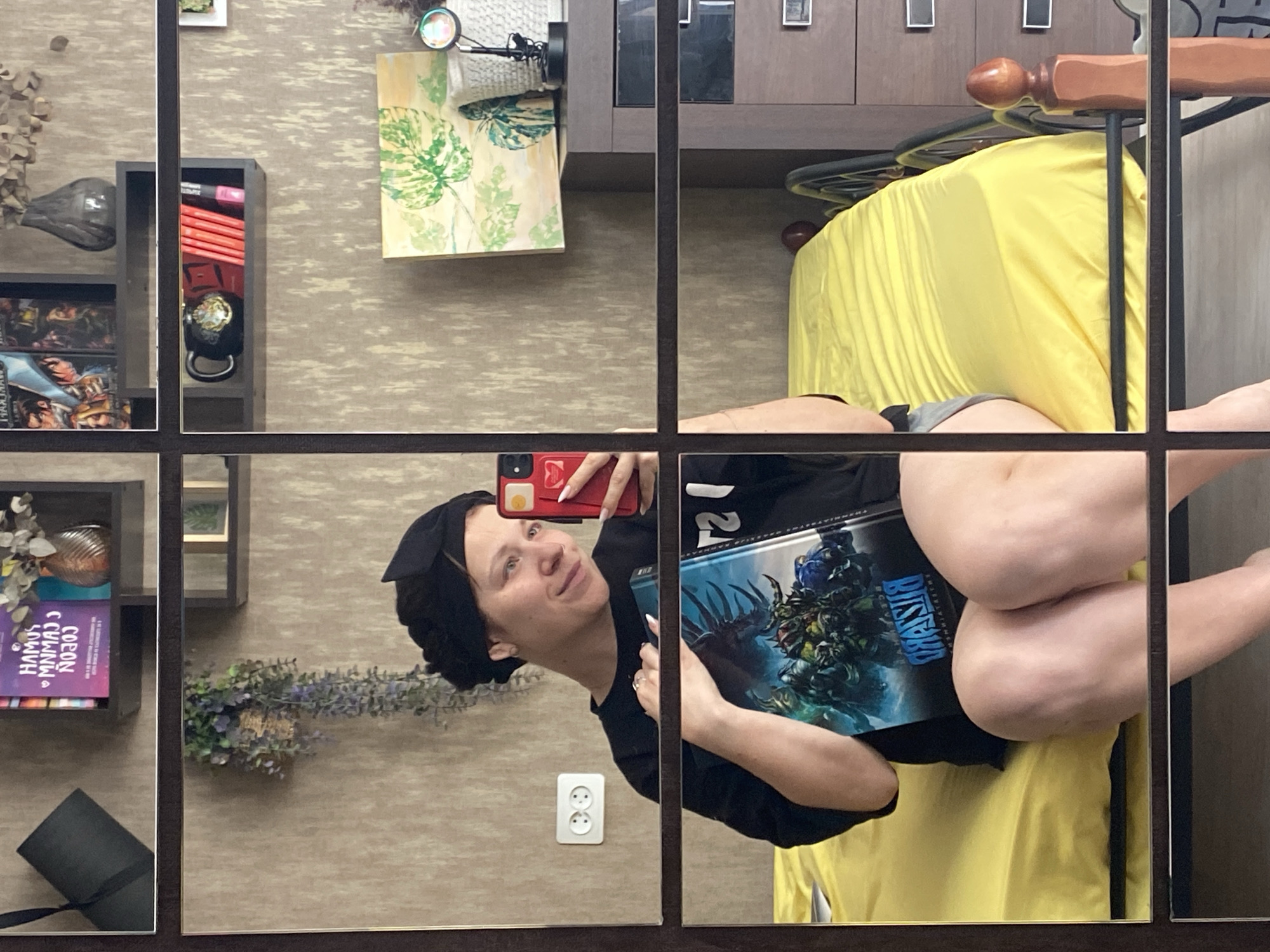 The image, presented rotated 90 degrees clockwise, depicts a mirror selfie taken by a woman in an indoor setting in Chaikovsky, Russia. The woman, wearing a black top, grey shorts, and a black head wrap, holds a red smartphone to capture the reflection and a large book or box displaying "BLIZZARD" branding alongside fantasy artwork. She has a pleasant, slightly smiling expression as she looks towards her phone.
The reflected room features textured, light brown wallpaper. To the woman's right (which would be behind her in an upright view), a bed with bright yellow bedding and a wooden headboard is visible. To her left, several dark, open shelves contain various items: books (with fragmented text like "HAMUS", "CONNAN", "MANMAJ", "NOCDN"), board games, a dark, bird-shaped decorative vase, and a kettlebell. A rolled-up black mat rests on a lower shelf, and a small arrangement of artificial lavender-colored flowers is placed nearby. Higher up on the wall, a painting featuring green leaves and a floor lamp with a colorful light effect are reflected. A white electrical outlet is also visible on the wall. The mirror itself is divided into a grid by dark vertical and horizontal frames. The scene suggests a casual, personalized bedroom or living space.