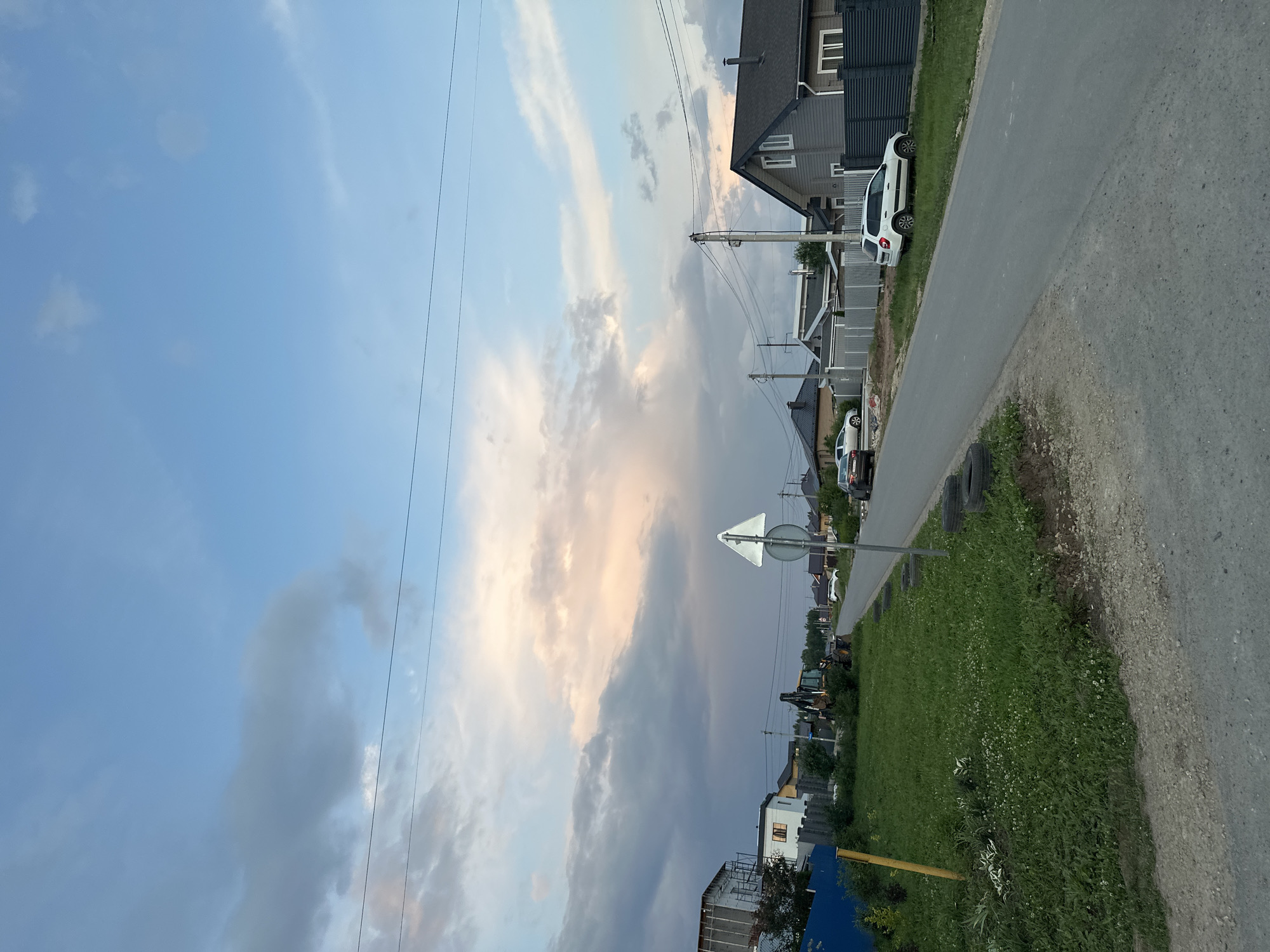The image depicts a residential street in the city of Estachi, Russia, at dusk. A paved road runs from the foreground into the distance, with houses and fences lining the right side. A white SUV is parked on the right side of the road. On the left side of the road, there is a grassy area with some vegetation and a yellow pole. A yellow backhoe is visible in the mid-ground on the left. The sky is partly cloudy with hues of pink and orange, indicating the setting sun. Power lines are visible across the upper portion of the image.