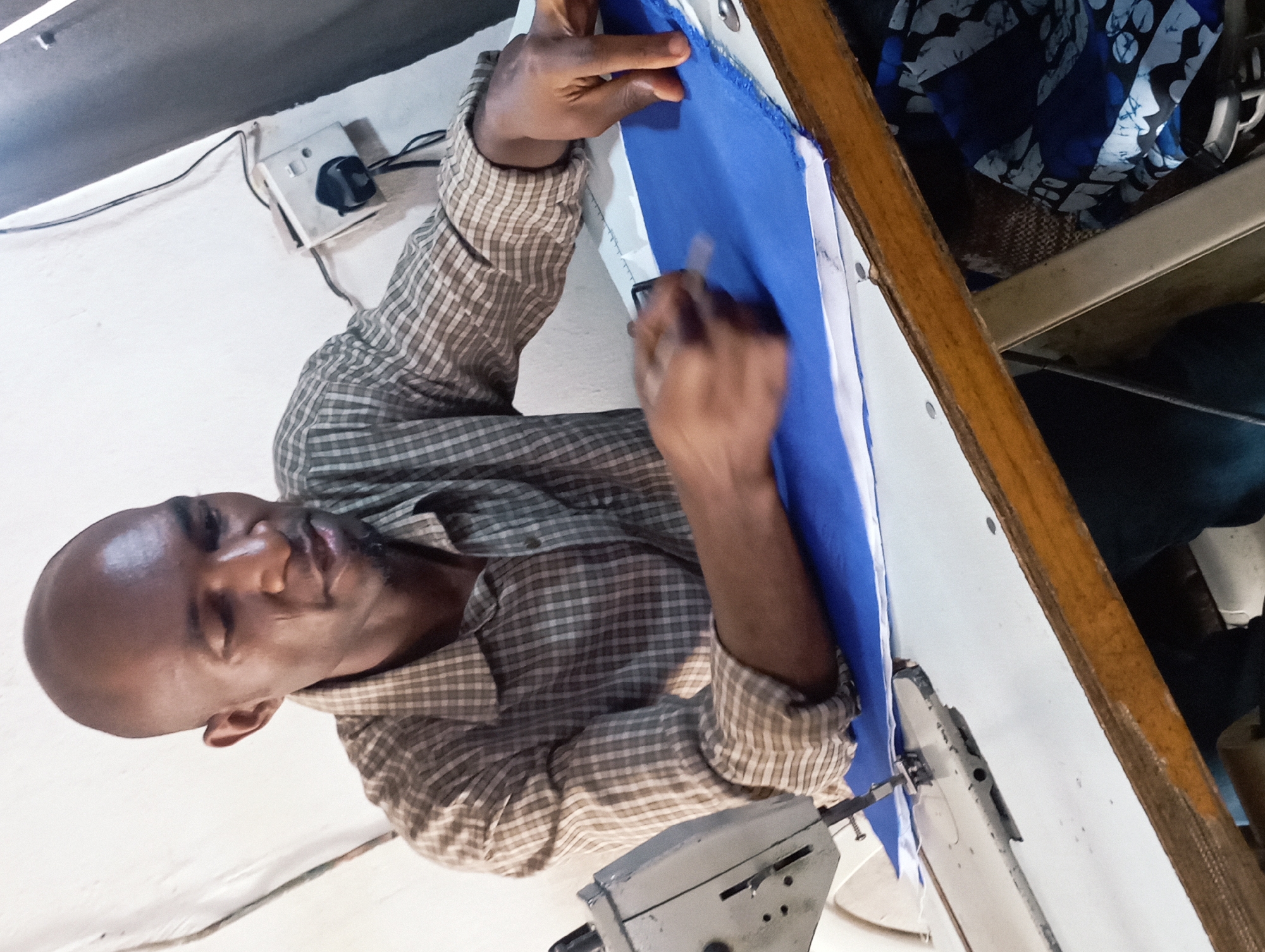 An African man is captured in a high-angle shot, intently focused on working with fabric at a sewing workstation in Jos, Nigeria. He has a bald or closely-shaven head and a light beard or stubble, and is dressed in a long-sleeved plaid shirt with a brown and white checkered pattern.
His hands are actively engaged with a vibrant blue piece of fabric, which appears to have a white lining or trim. His left hand gently holds the fabric in place, while his right hand, slightly blurred from motion, holds a small, light-colored tool, possibly for marking or guiding the fabric. A portion of a sewing machine, identifiable by its metallic base and needle area, is visible in the lower foreground, indicating the nature of his craft.
The setting is an indoor workshop environment, characterized by a plain white wall in the background. An electrical outlet or switch with a dark knob and associated wiring is mounted on the wall. To the right, behind the man, other textiles are visible, including a blue and white patterned fabric, possibly with a batik design, along with elements of wooden and metal shelving. The man's expression conveys concentration and diligence as he works. The lighting suggests it is daytime, and the overall scene depicts a skilled individual engaged in tailoring or textile work. No discernible text is visible within the image.