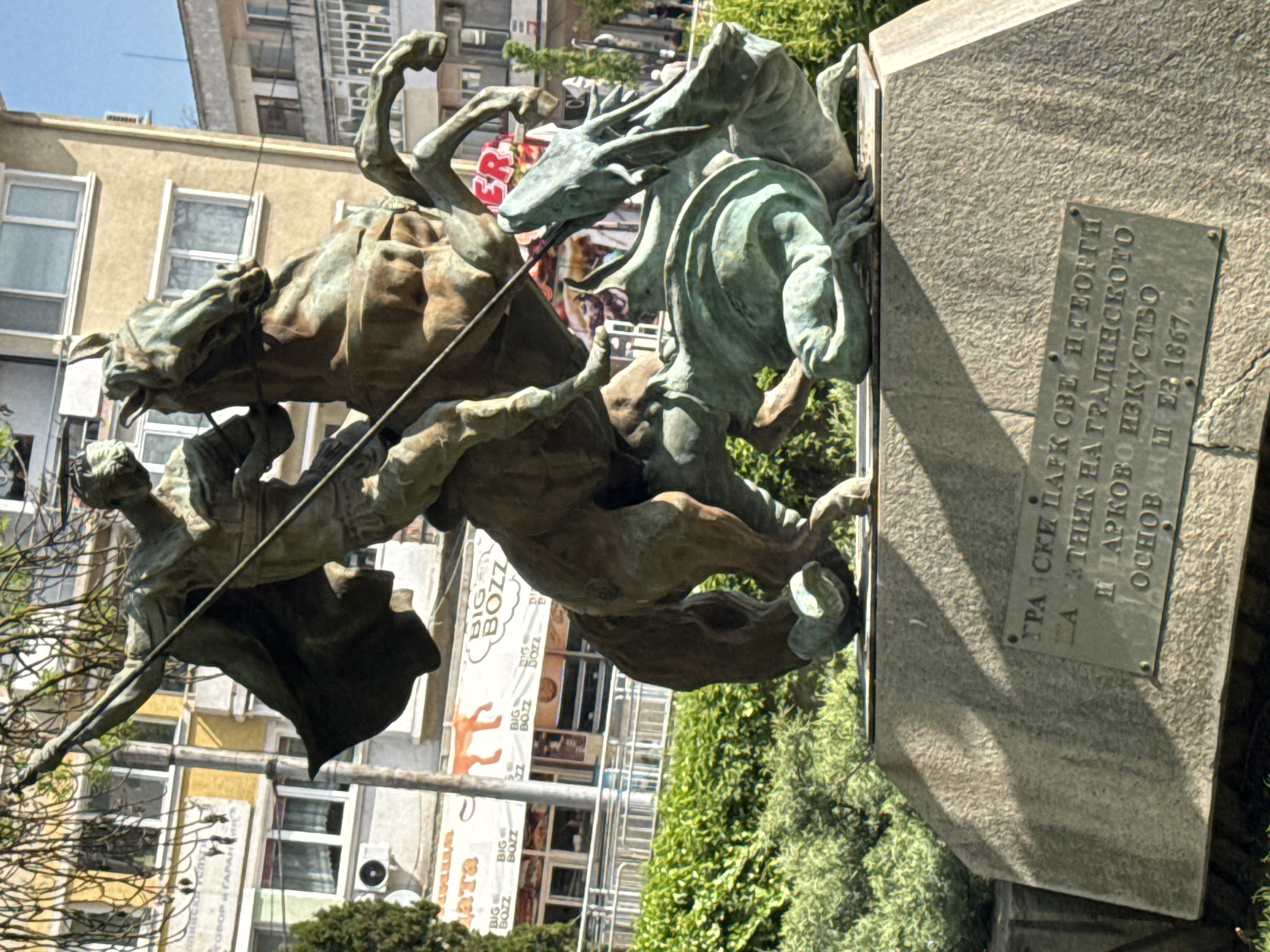 The image captures a bronze statue of Saint George slaying a dragon. The statue, featuring a man on horseback battling a dragon, is mounted on a gray stone base. The base includes a bronze plaque with Cyrillic inscriptions. In the background, buildings are visible, including one with signage for "BIG BOZZ". The statue is surrounded by greenery. The scene appears to be in a public park or square in Dobrich, Bulgaria, judging by the Cyrillic script and the typical urban environment.