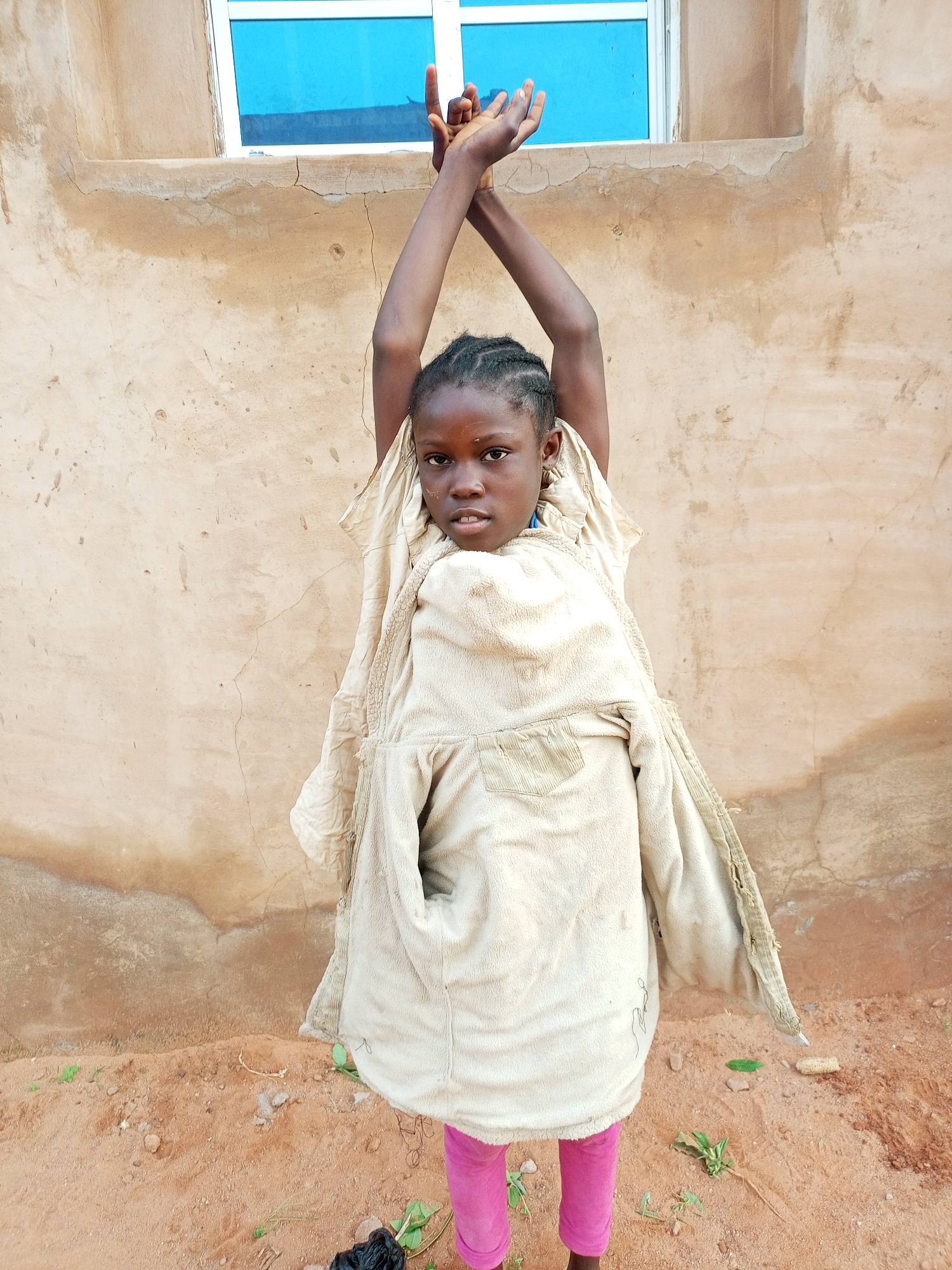 A young girl stands in front of a textured, light brown wall with a blue-windowed building above. She is wearing a cream-colored, fleece-lined jacket over pink leggings. Her arms are raised above her head, with her hands clasped. Her hair is braided and pulled back from her face. The ground in front of her is sandy dirt with a few small green plants scattered about. The overall scene appears to be outdoors, possibly in a residential area of Tempure, Nigeria, given the wall construction and window style.