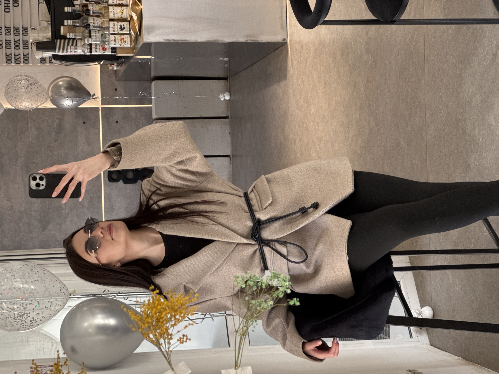 A woman wearing sunglasses, a beige coat with a black drawstring, and black leggings is taking a selfie. She is holding a smartphone in her left hand. Two balloons, one silver and one clear with silver glitter, are visible in the lower left. In the bottom center, there are two small vases, each with a different type of dried flower.
In the background, various bottles and containers are placed on a shelf. Above these, text is readable, including the words "SY BICHESY," "BICHESY," and "OHA 100 180 500," which likely relate to the city of Chaikovsky, Russia. A wall and floor, both in neutral tones, make up the rest of the background.
