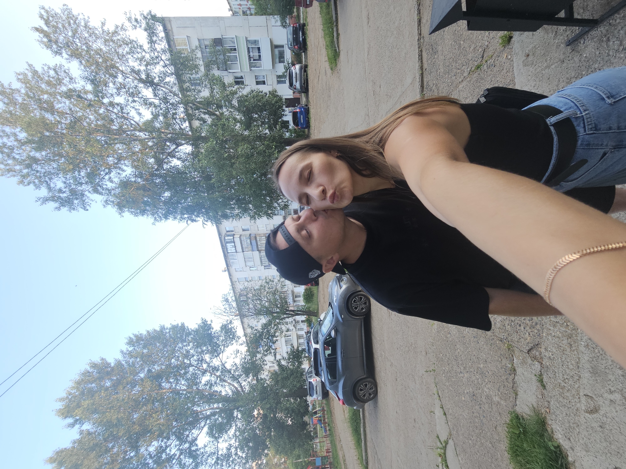 The media file shows two young adults, a man and a woman, in an outdoor urban setting. The man is wearing a black t-shirt and a black baseball cap. The woman has long brown hair and is wearing a black top and blue jeans. They are in the process of sharing a kiss, with the woman leaning in towards the man and puckering her lips. The scene appears to be a residential area, with apartment buildings and parked cars visible in the background. Trees with green foliage suggest it is daytime, likely during warmer months, and the sky is a clear blue. The overall mood appears affectionate and romantic. There is no visible text in the image.