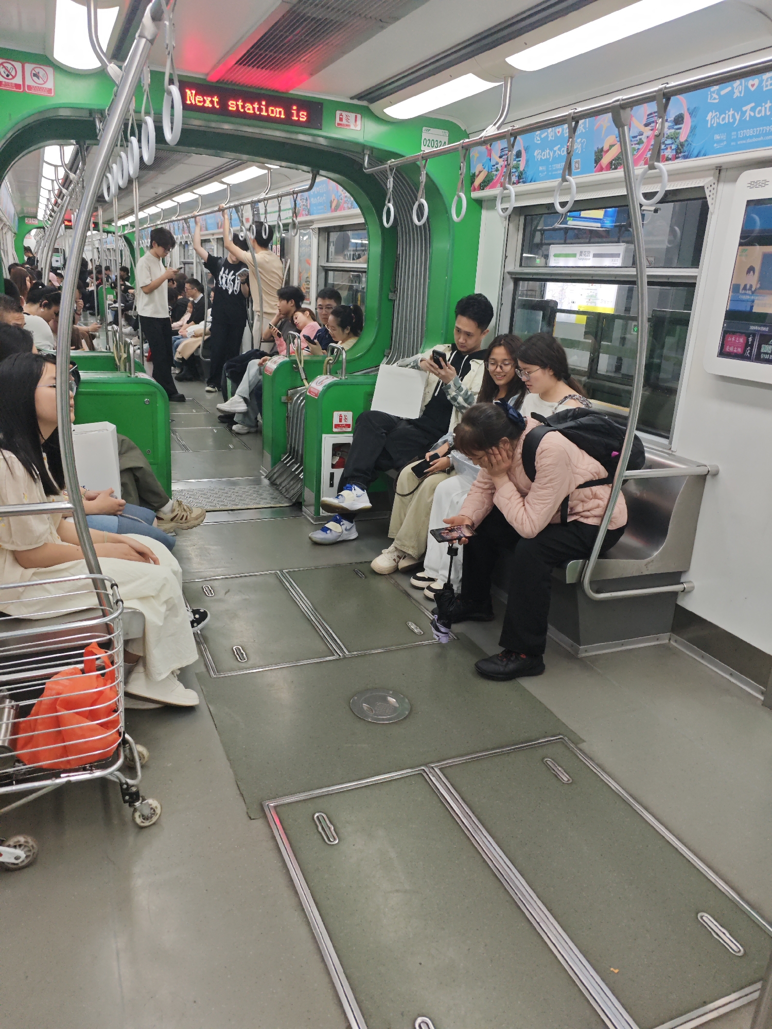 The image captures a scene inside a subway car. Multiple passengers are present, some seated and others standing, occupying the available spaces. Many passengers are engaged with their mobile phones.
A text display indicates "Next station is" in English. There are also advertisements in Chinese characters on the walls. The interior design includes green color accents.
The advertisements contain phone numbers starting with 137, suggesting a location in China. Given the other details and the prevalence of Chinese text, it is highly probable that this scene is from a subway in a Chinese city. The green interior design and signage match the Yuzhong District, China subway system.