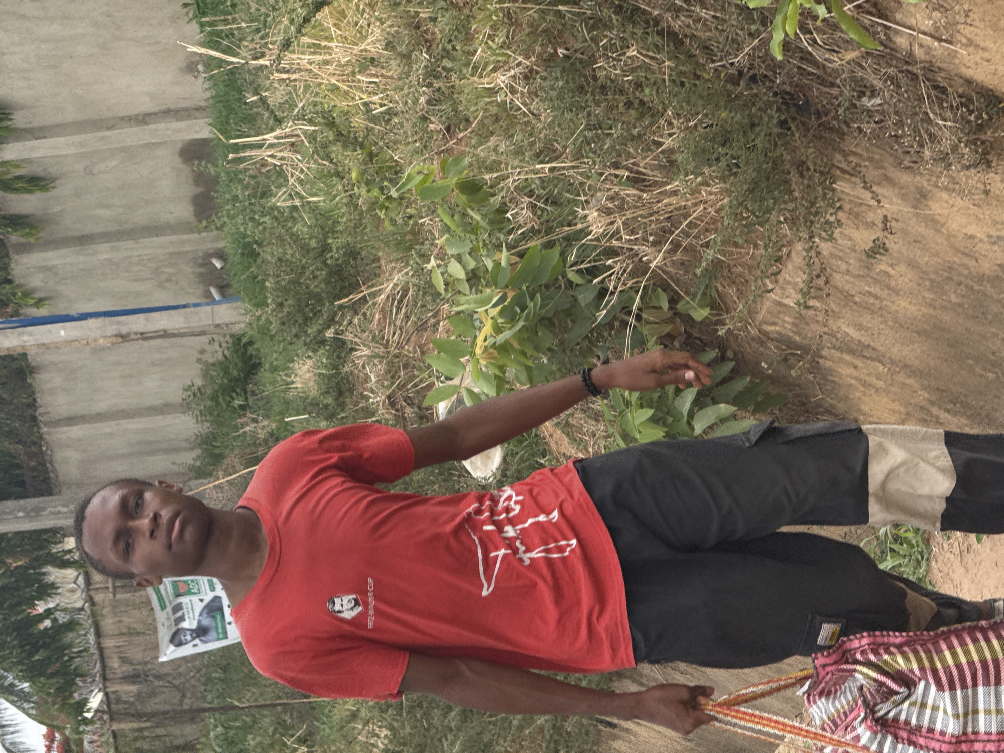 A young man stands in an outdoor setting that appears to be on the periphery of a built-up area, potentially in Abuja, Nigeria. He is wearing a red t-shirt with a white design, dark-colored cargo pants with a beige stripe, and a black bracelet on his left wrist. He is holding a striped bag with orange-ish handles. The background includes a concrete wall, shrubs, and dry grassy areas. A political poster is visible behind him. The ground appears to be a mix of soil and vegetation. The lighting suggests it is daytime.