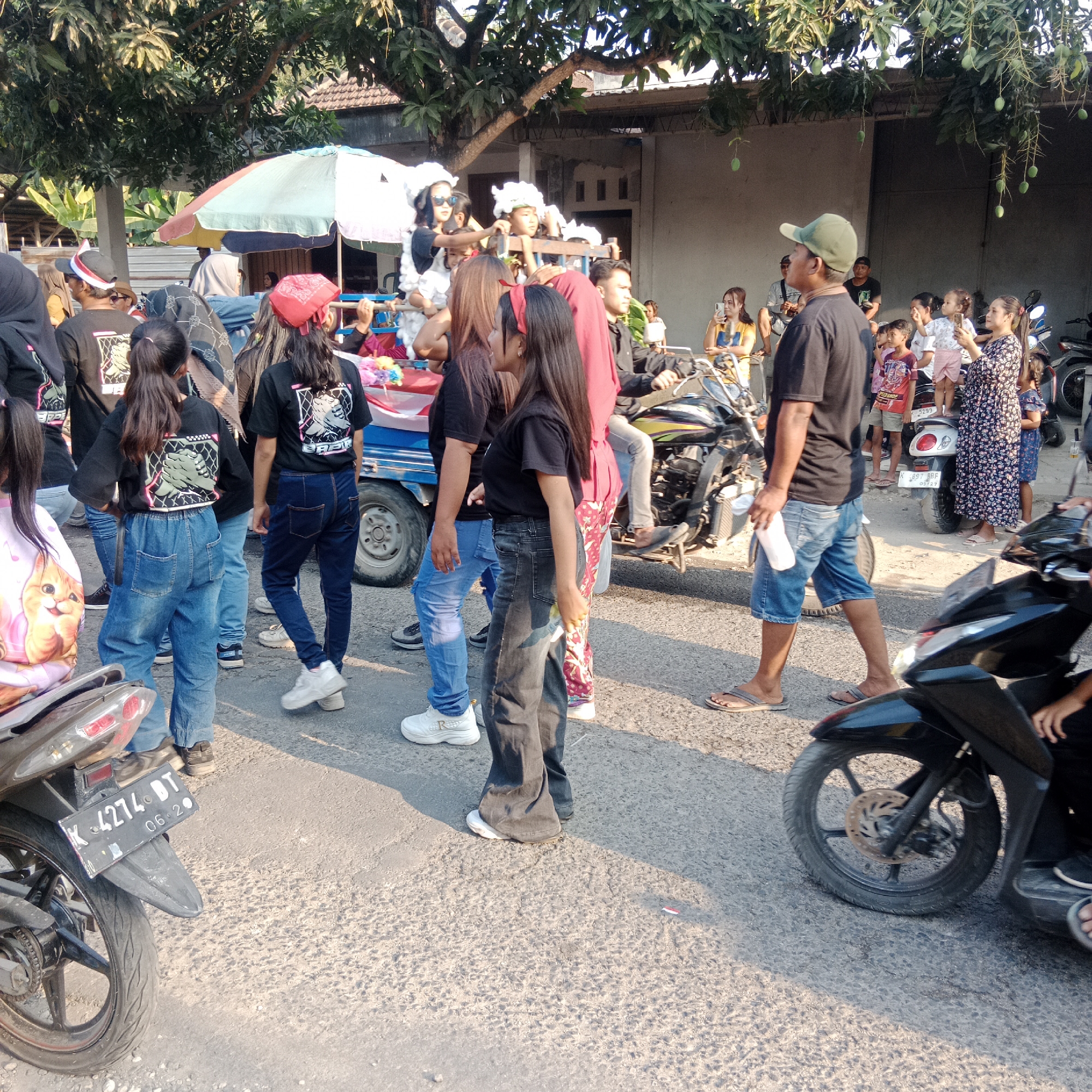 In Tegalorejo, Indonesia, a group of people, predominantly young women, are participating in a parade. They are dressed in black t-shirts with matching graphic designs on the back, and various styles of jeans. Some individuals are adorned with red headbands. A small blue truck, decorated with colorful flowers and possibly a banner, is visible, suggesting it is part of the parade float. Several motorcycles are also present, with some people riding on them and others standing around. The event appears to be taking place outdoors, on a paved road, under the shade of trees laden with mangoes. The atmosphere seems festive, with people engaging with each other and observing the parade. The time of day is likely late afternoon, given the long shadows cast by the trees and the warm lighting.