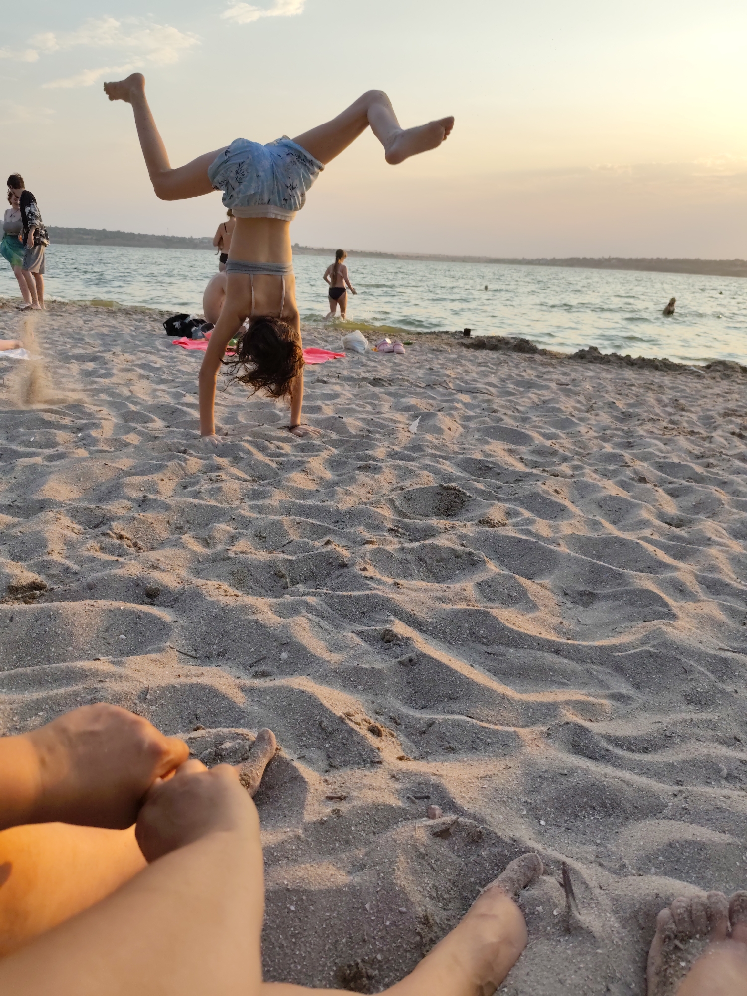 A young girl is performing a handstand on a sandy beach with the sea in the background. She is wearing blue shorts and a light-colored bikini top. Her legs are extended upwards in the air, and her hair is falling around her face. In the distance, several other people are visible, some in the water and some on the shore. The sun is setting, casting a warm, golden light on the scene. The sky is a soft pink and orange, and the clouds are scattered. The waves are gentle, and the water is calm. The beach is covered in sand, with some scattered debris like twigs and shells. The overall mood of the image is peaceful and relaxed, with a sense of fun and playfulness. The location appears to be a coastal area in Mykolaiv, Ukraine, given the context.